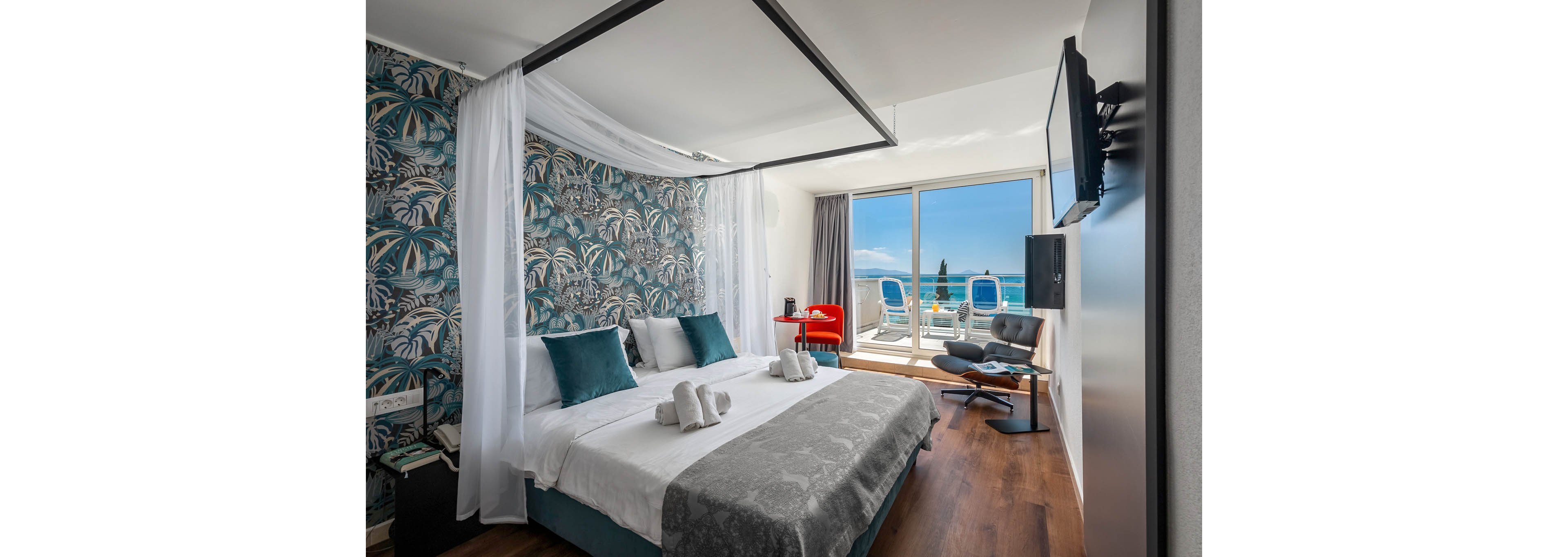 A contemporary hotel room features a canopy bed with teal accent pillows and a tropical-patterned wall. The space includes a seating area with a red chair and a lounge chair, facing a large window that opens to a balcony overlooking the ocean. The room is decorated in cool tones, with wood flooring and a flat-screen TV mounted on the wall. Two white towels are arranged on the bed, and the balcony has two blue and white chairs.