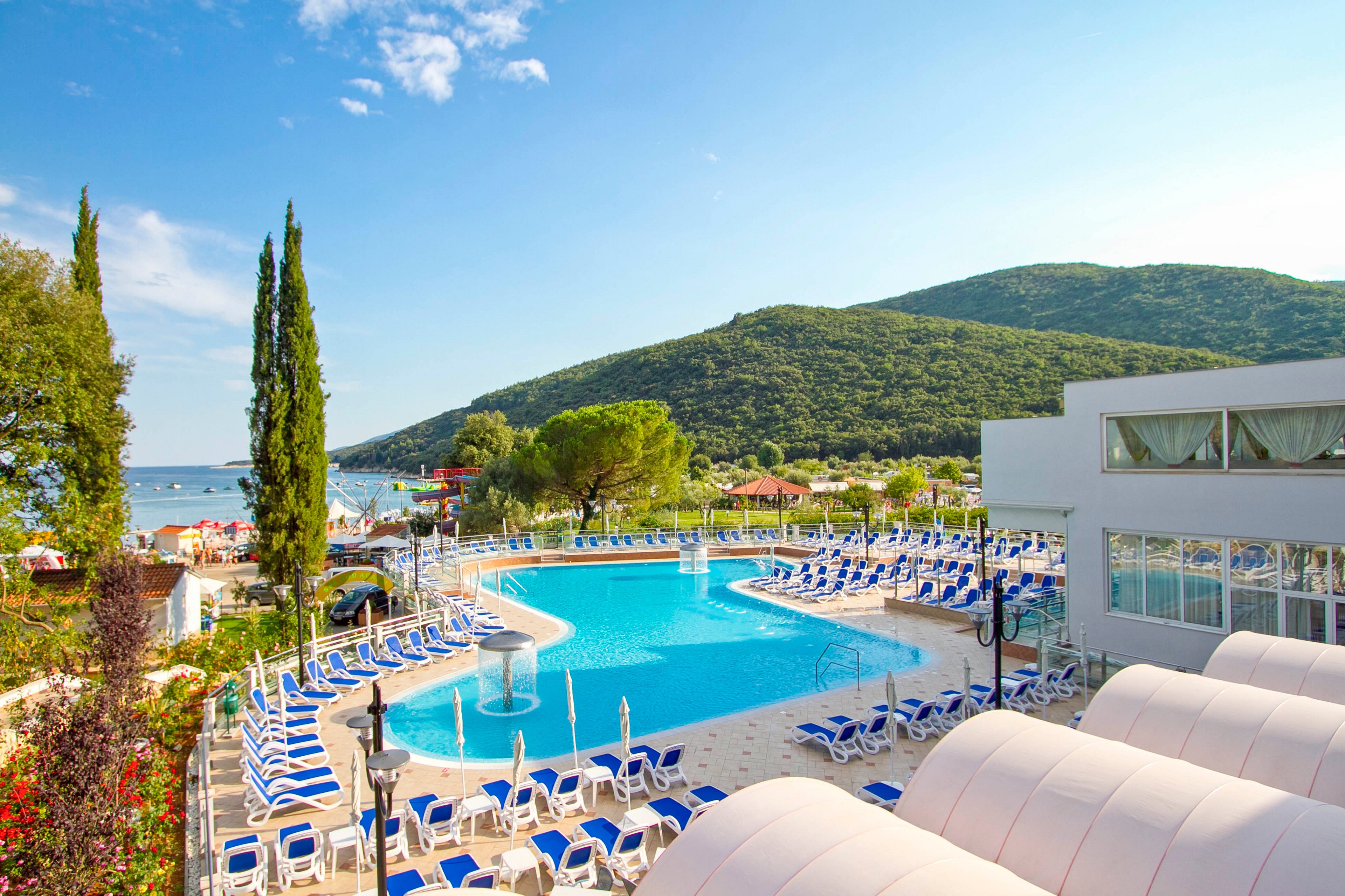 A large outdoor swimming pool is surrounded by numerous blue and white lounge chairs at a modern resort. The setting features lush green hills and a glimpse of the sea in the background, creating a relaxing vacation atmosphere. The scene is bright and sunny, with clear skies and vibrant colors. No visible people or text are present in the image.
