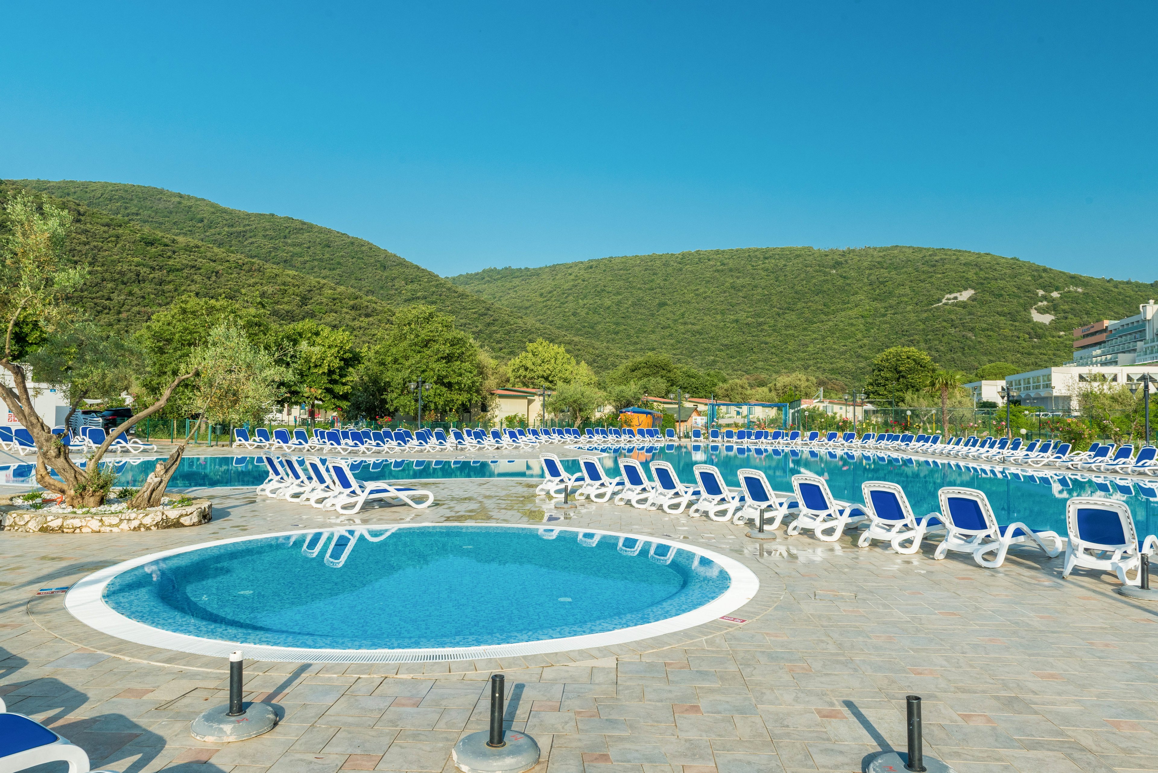 A large outdoor swimming pool area is surrounded by numerous white and blue lounge chairs, set against a backdrop of lush green hills. The scene features a smaller circular pool in the foreground and a spacious main pool behind it. The setting is bright and sunny, evoking a relaxing vacation atmosphere. No people are present, and the sky is clear blue, enhancing the tranquil mood.