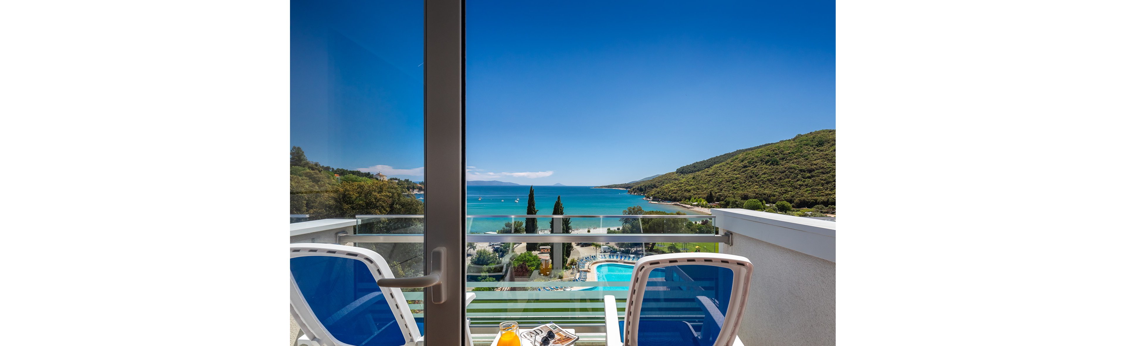 A bright balcony scene features two blue and white chairs facing a stunning turquoise sea and a swimming pool below. The setting is a sunny coastal resort with lush green hills and clear blue skies. On the table, a glass of orange juice and a book suggest a relaxing atmosphere. The overall mood is tranquil and inviting, perfect for a summer getaway.