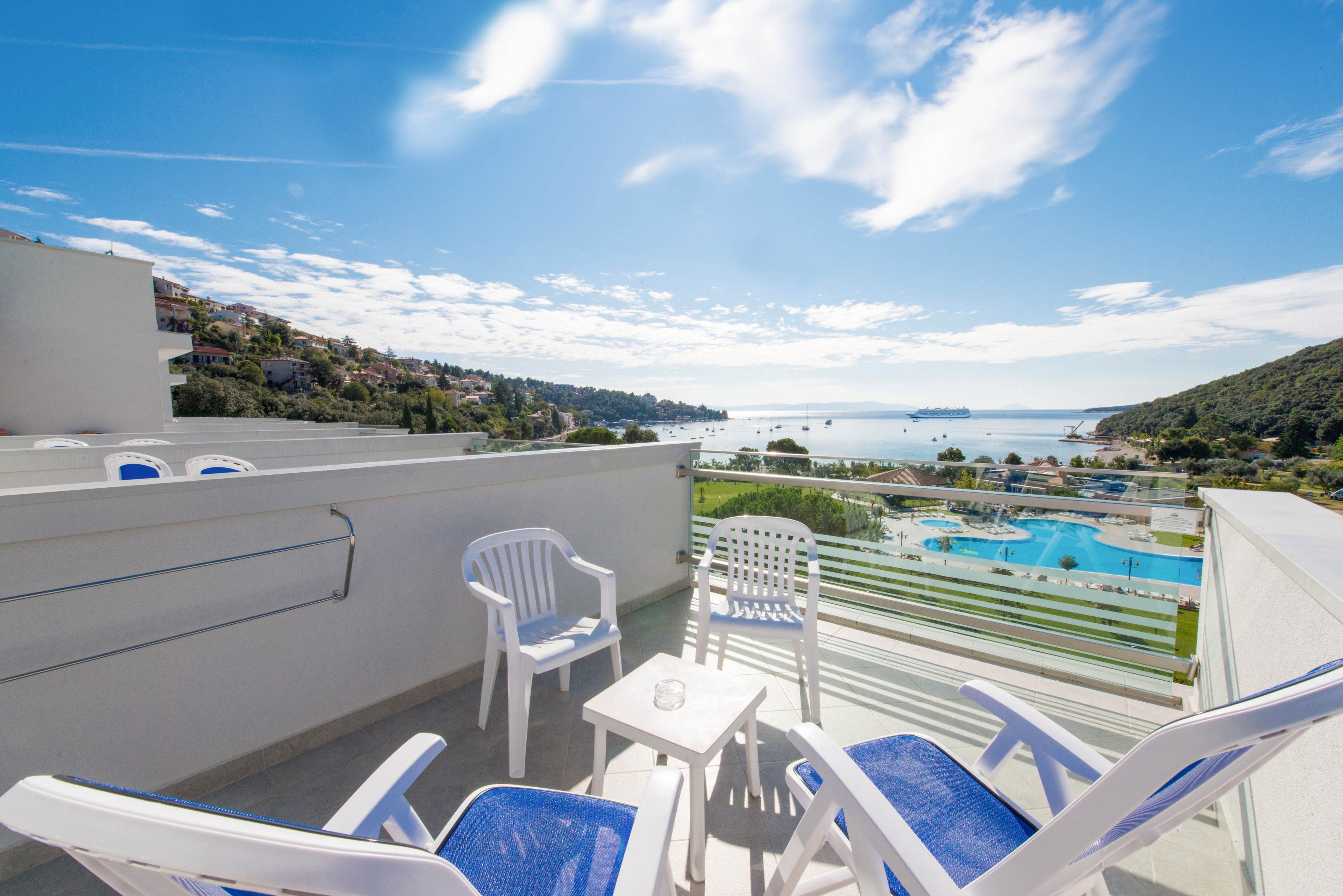 A bright outdoor terrace features four white plastic chairs and a small table, overlooking a scenic coastal landscape. The setting includes a clear blue sky, distant hills, and a sparkling swimming pool below, with the sea visible on the horizon. The mood is relaxed and inviting, ideal for leisure or vacation. The color palette is dominated by whites and blues, enhancing the fresh, summery atmosphere.