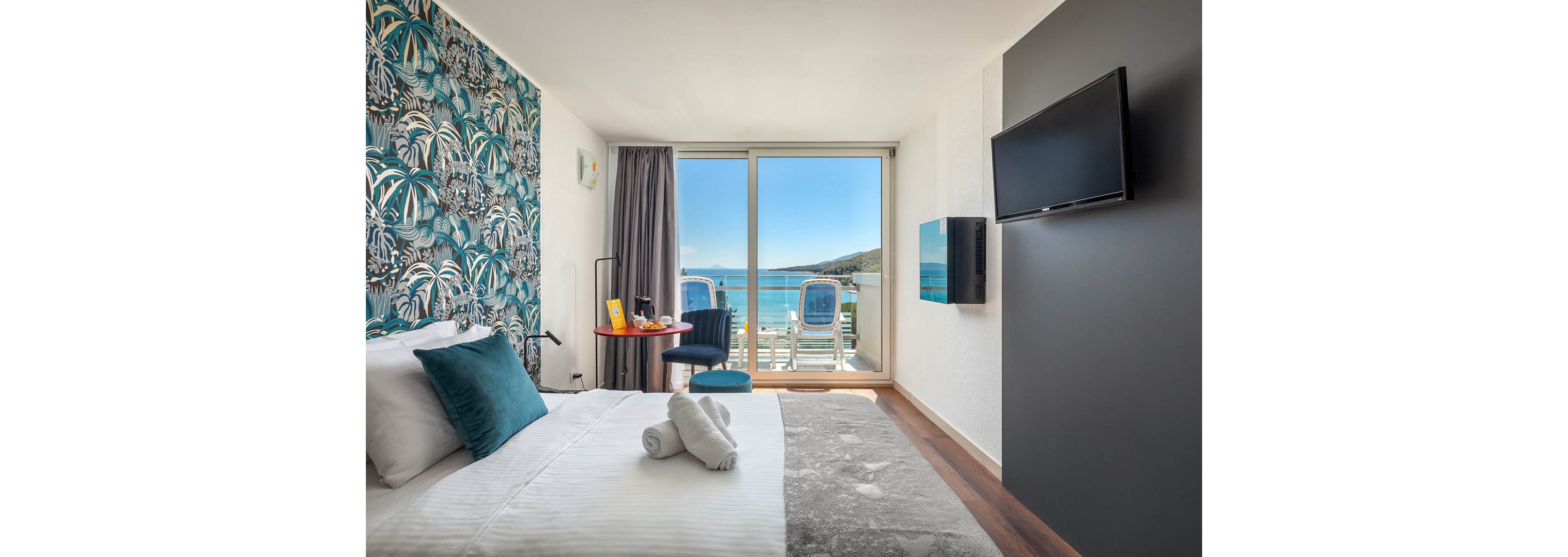 A contemporary hotel room features a neatly made bed with white linens and teal accents, facing a wall-mounted television. The room opens to a balcony with two chairs overlooking a bright blue sea and distant hills. Tropical-patterned wallpaper adds a stylish touch, while natural light fills the space, creating a fresh and inviting atmosphere. No visible text or numbers are present in the image.