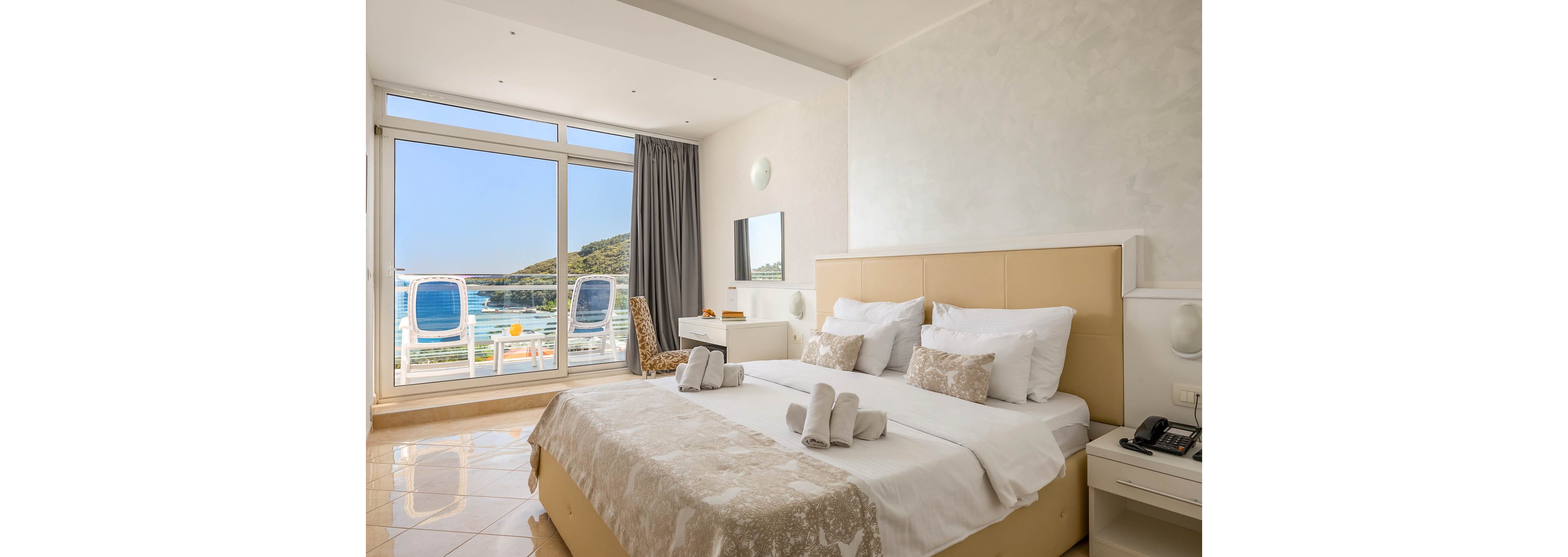 A bright, contemporary hotel room features a large bed with neatly arranged towels and pillows. The space opens to a balcony with two blue lounge chairs overlooking a scenic waterfront and hills. Neutral tones and natural light create a calm, inviting atmosphere. A desk and chair are positioned near the window, enhancing the room's functionality. The overall mood is serene and relaxing, ideal for a vacation stay.