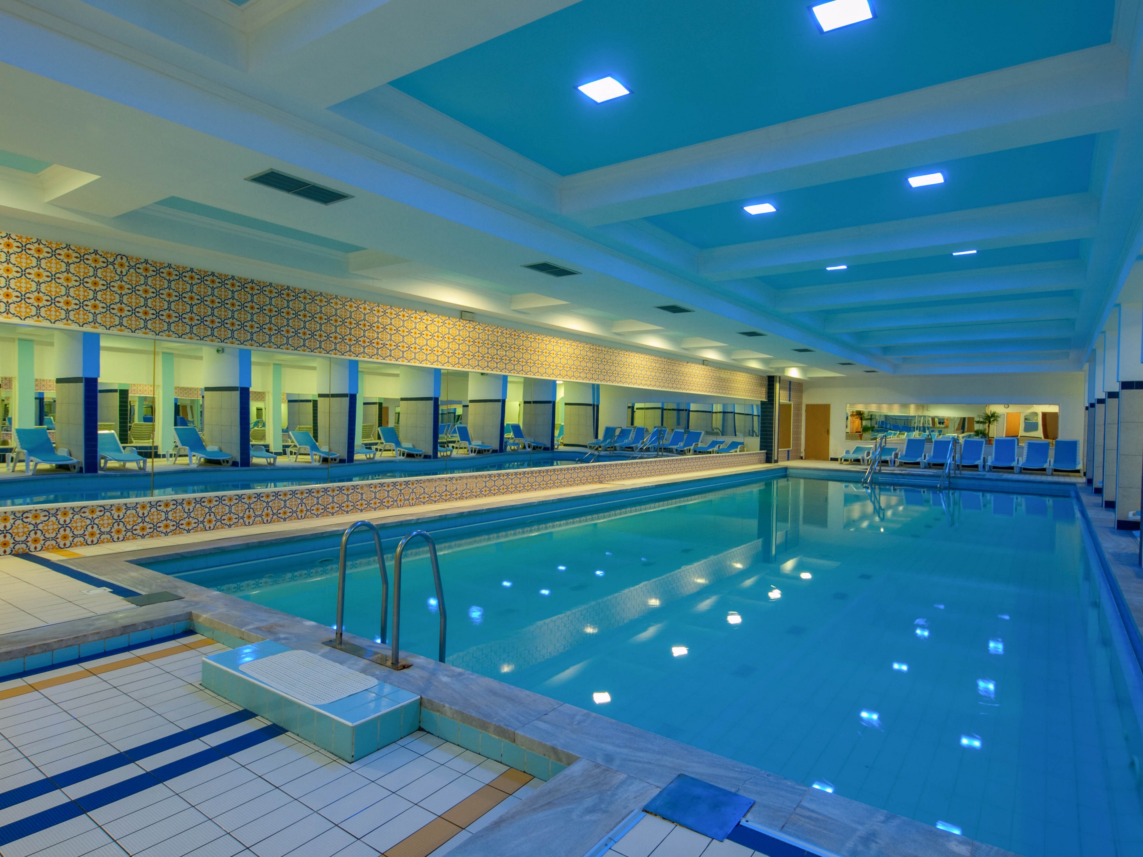 A spacious indoor swimming pool is featured in this image, surrounded by blue lounge chairs and decorative tiled walls. The pool area is well-lit with ceiling lights, and large mirrors enhance the sense of space. The setting appears clean and inviting, with a cool blue and white color palette. No people are present, emphasizing a calm and serene atmosphere.