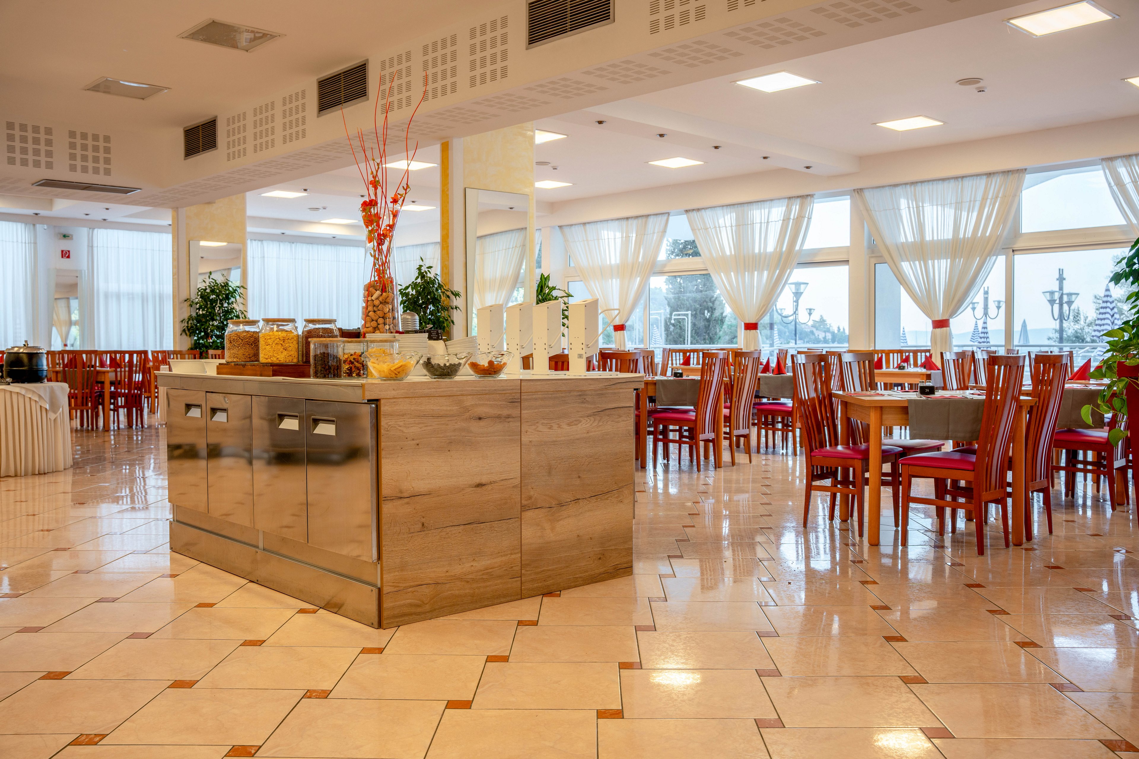 A spacious, well-lit hotel dining room features a central wooden buffet station with jars of cereals and snacks. The setting includes polished tile floors, large windows with sheer curtains, and neatly arranged wooden chairs around tables. Decorative plants and minimalist vases add to the contemporary ambiance. The room is empty, emphasizing a clean and inviting atmosphere.