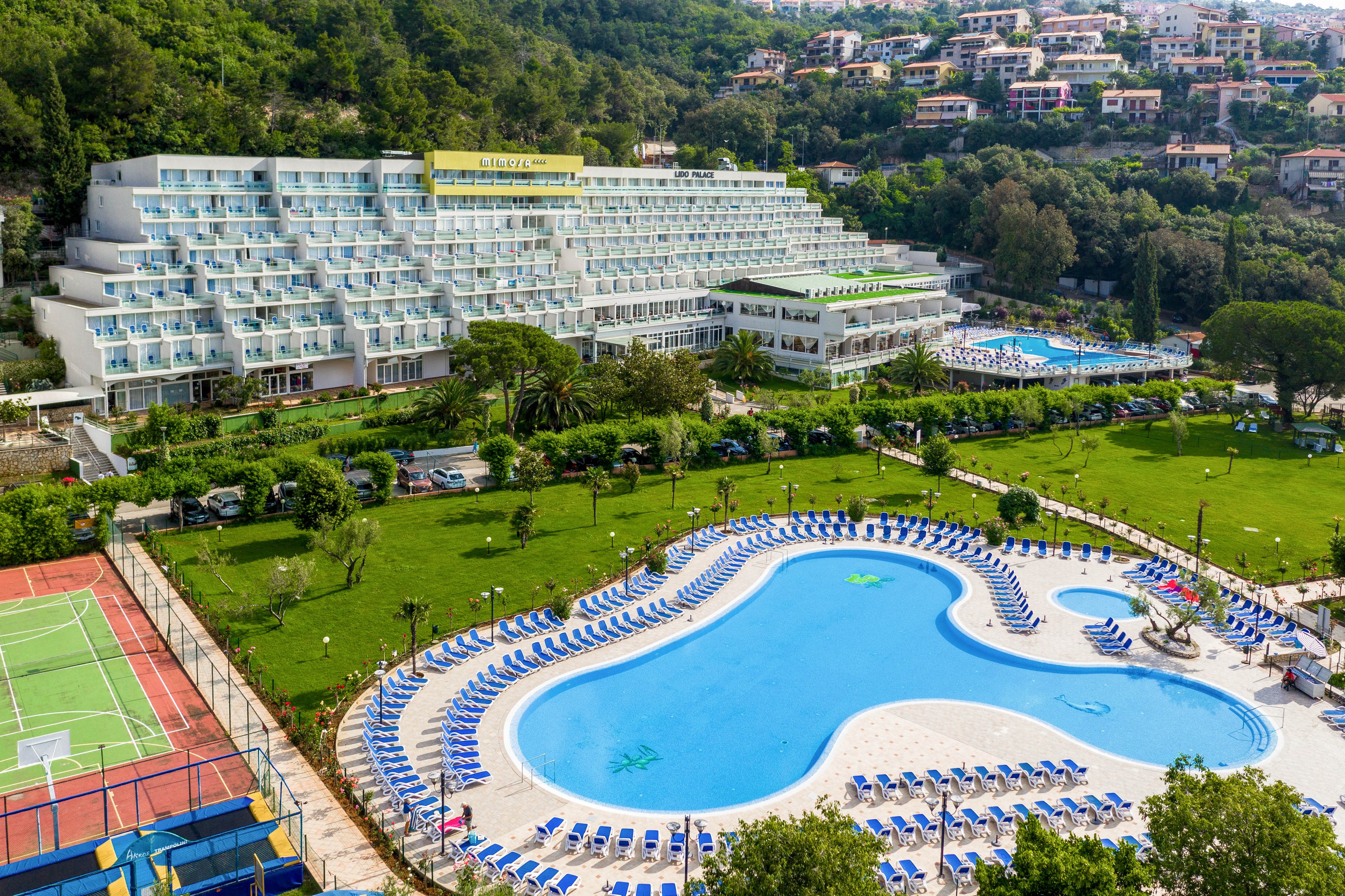 A multi-story resort hotel is set against a lush hillside, featuring a large, uniquely shaped outdoor swimming pool surrounded by numerous blue lounge chairs. The property includes a tennis court and expansive green lawns, with additional pools visible in the background. The setting is vibrant and inviting, ideal for leisure and recreation, with clear signage reading 'GRAND HOTEL' on the building.