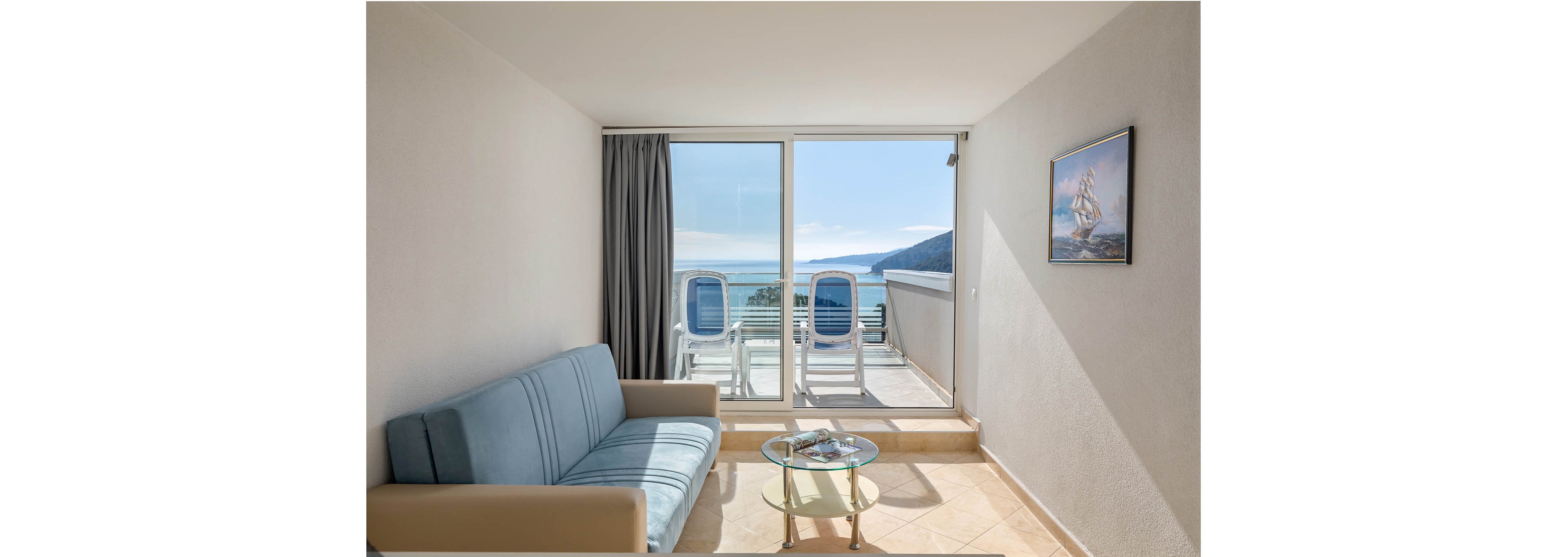 A bright, minimalist hotel room features a light blue sofa and a glass coffee table with magazines. Large sliding glass doors open to a balcony with two white lounge chairs, overlooking a serene blue sea and distant hills. The room is decorated with a nautical-themed painting and has a neutral color palette, creating a calm and inviting atmosphere.