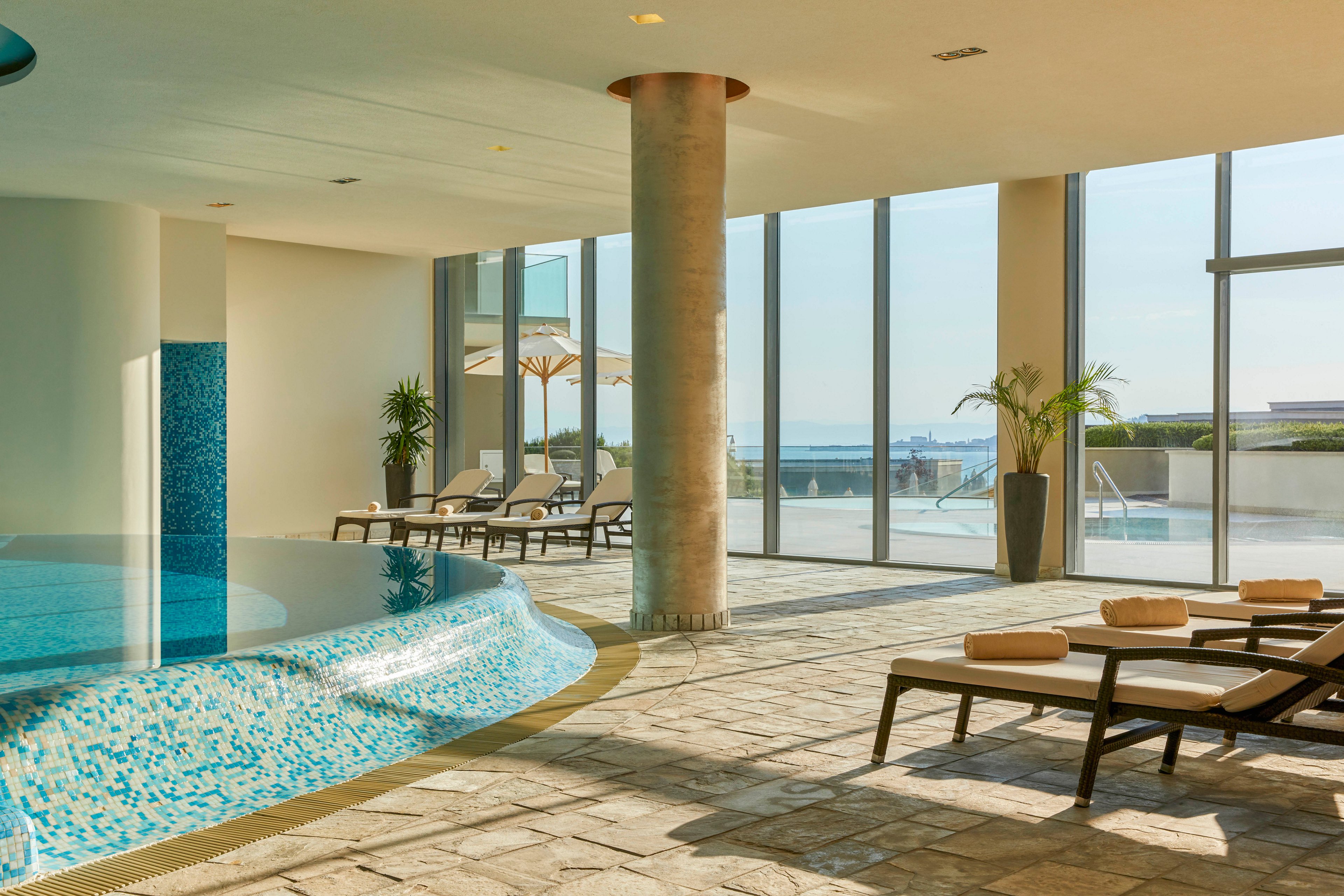 A contemporary indoor pool with mosaic tiles is featured in a spacious, sunlit room. Several cushioned loungers with rolled towels are arranged near large floor-to-ceiling windows, offering a view of an outdoor pool and distant water. The setting is tranquil and luxurious, with neutral tones and natural light enhancing the relaxing atmosphere. Potted plants add a touch of greenery to the serene environment.