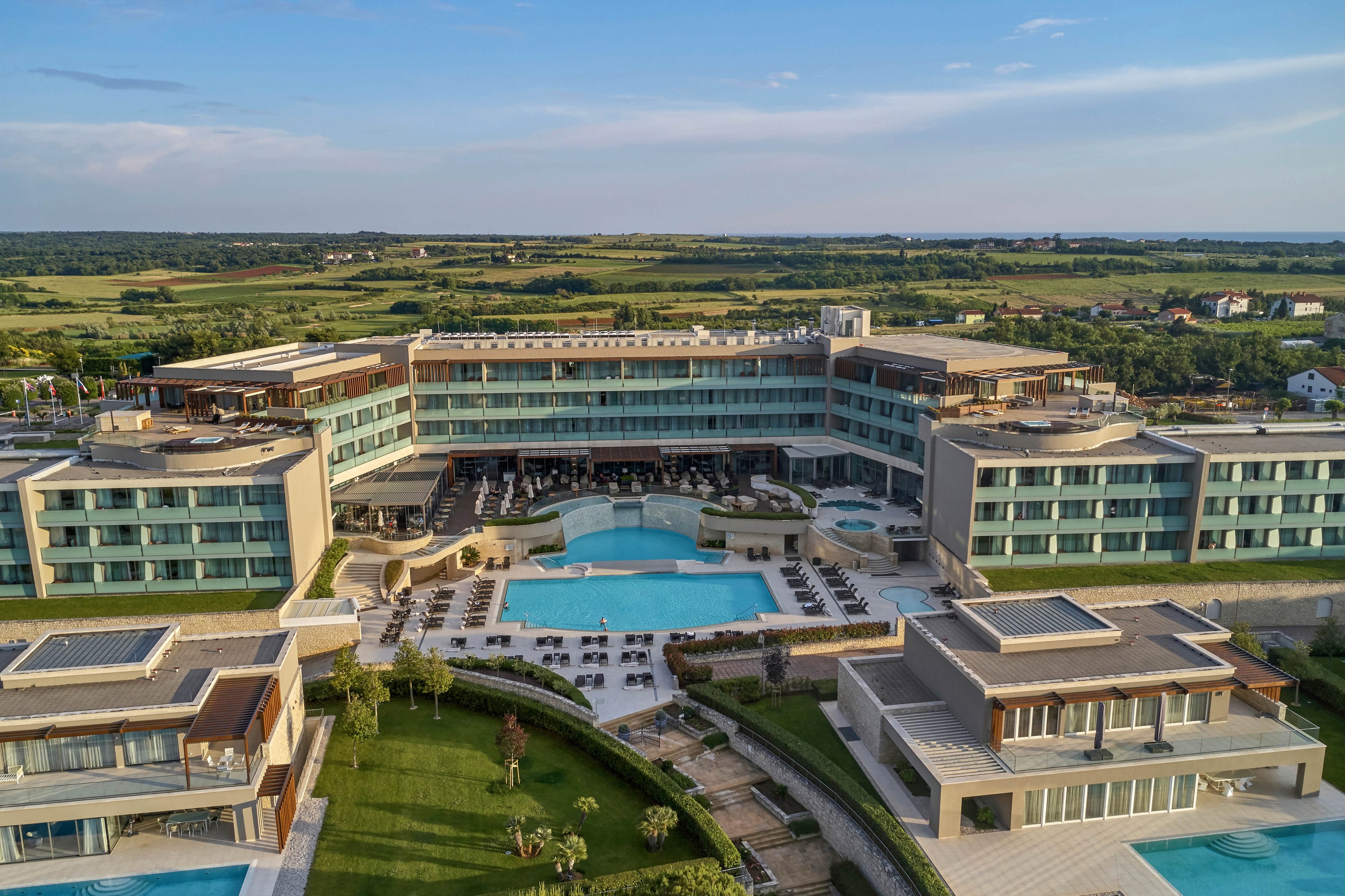 Aerial view of a contemporary resort complex featuring several outdoor swimming pools and sun loungers. The architecture is modern with clean lines and large glass windows, surrounded by manicured lawns and landscaped gardens. The setting is tranquil, with open countryside visible in the background under a clear sky. The overall mood is relaxed and inviting, ideal for leisure and vacation stays.