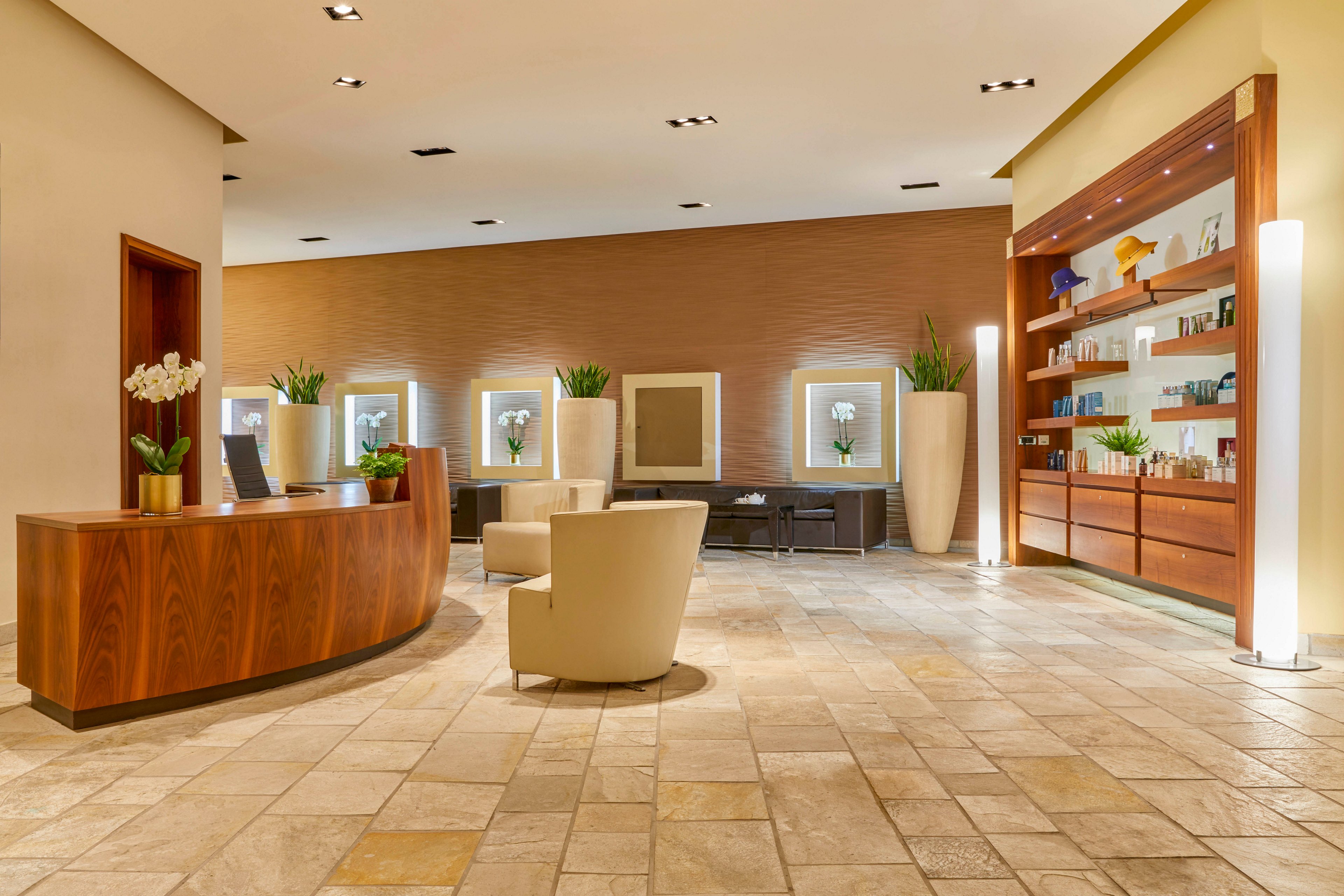 A contemporary spa reception area features a curved wooden desk, comfortable beige chairs, and large decorative planters. The setting is bright and welcoming, with warm lighting and a neutral color palette. Shelves display various products, and the tiled floor adds a touch of sophistication. The overall mood is calm and inviting, ideal for relaxation and wellness. No visible text or numbers are present in the image.
