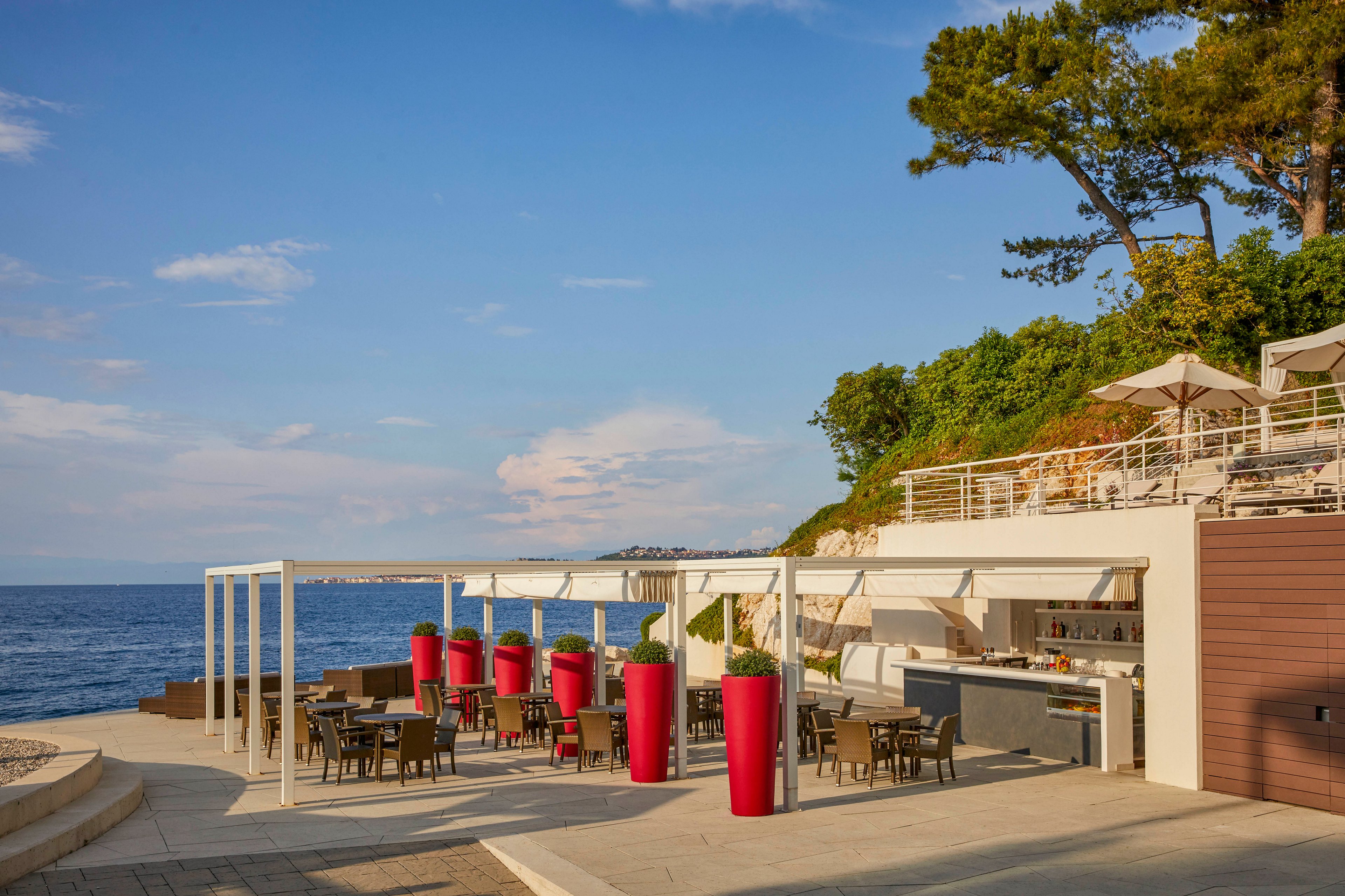 A contemporary outdoor restaurant is set along a scenic coastline, featuring tables and chairs under a white pergola. Large red planters add a pop of color to the neutral palette, while the open-air bar and dining area overlook the calm blue sea. The setting is tranquil and inviting, with lush greenery and umbrellas on an upper terrace enhancing the relaxed, vacation atmosphere.