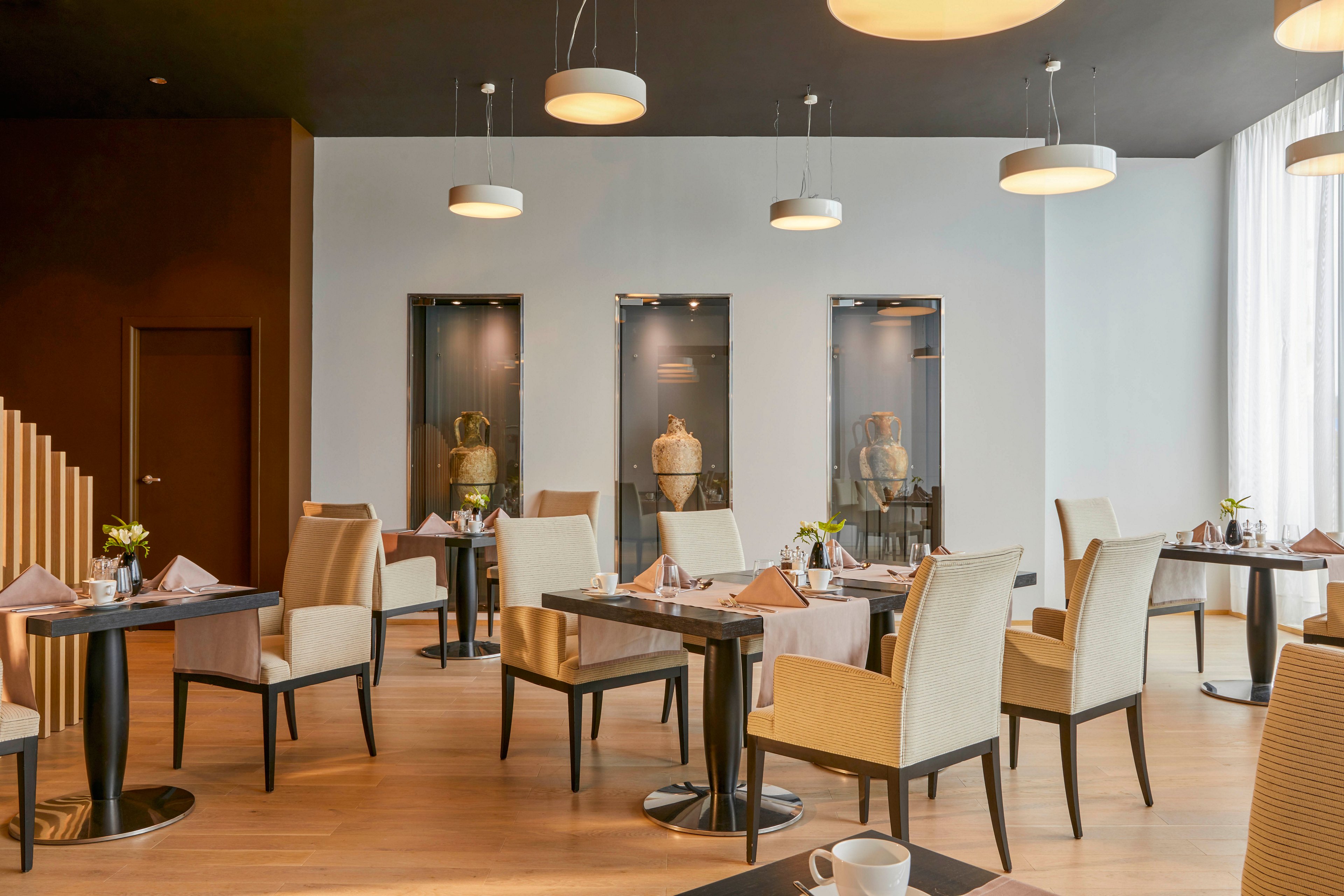 A contemporary restaurant dining area features neatly arranged tables and cushioned chairs. The setting is bright with natural light from large windows and modern pendant lighting overhead. Decorative vases are displayed in wall niches, adding a sophisticated touch. The overall mood is calm and inviting, with neutral tones and minimalist decor. No visible text or numbers are present in the image.