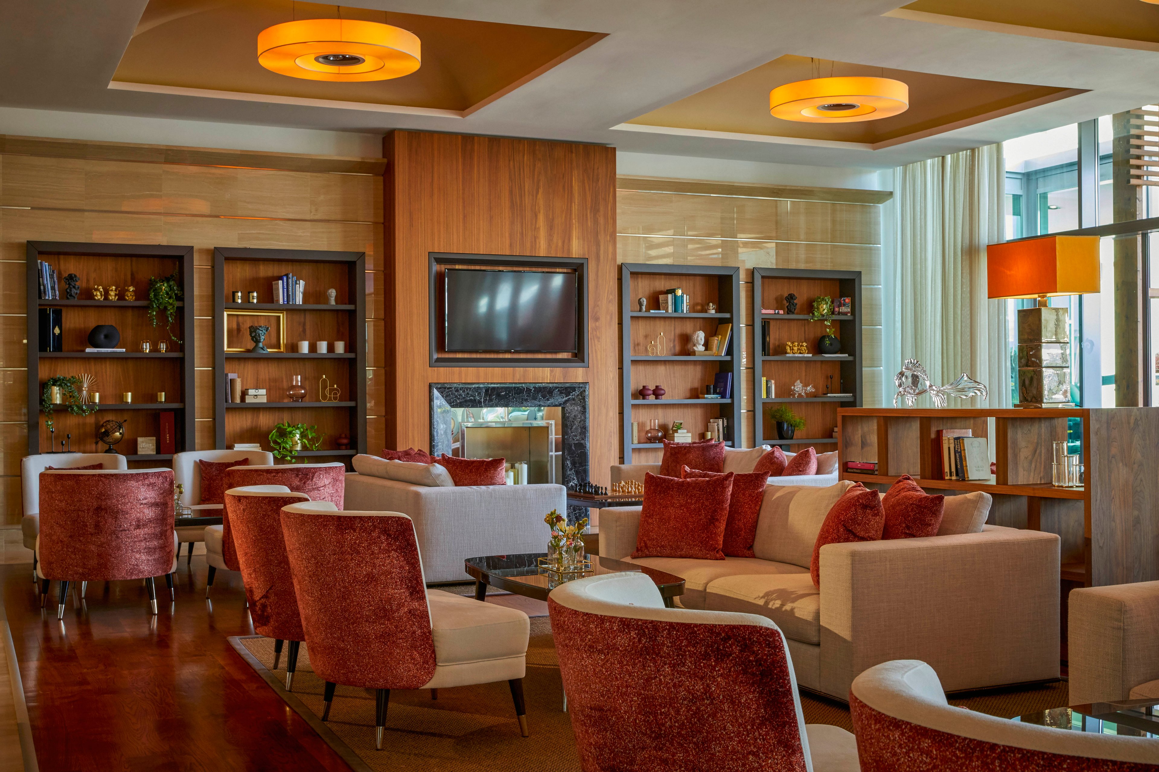 A contemporary hotel lounge features plush armchairs and sofas arranged around a central fireplace and television. The setting is warm and inviting, with wooden bookshelves, decorative objects, and soft lighting from ceiling fixtures. The color palette includes earthy tones, beige, and deep red accents, creating a cozy and sophisticated atmosphere. Large windows allow natural light to fill the space, enhancing the modern interior design.