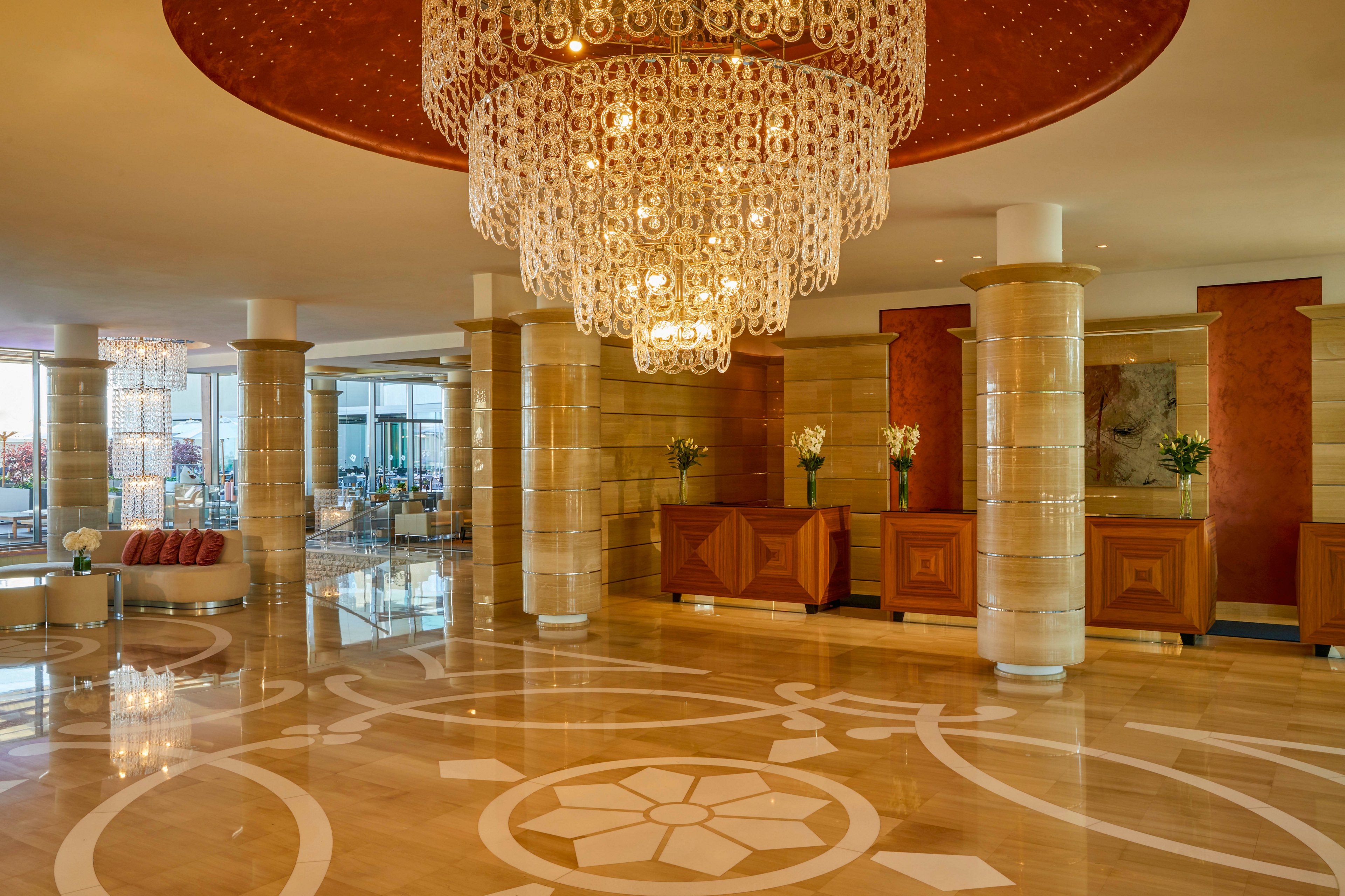 A spacious hotel lobby features a large, ornate chandelier as its centerpiece, casting warm light across polished marble floors with intricate patterns. The setting includes tall columns, a reception desk with flower arrangements, and modern decor elements. The overall mood is luxurious and welcoming, with a neutral and gold color palette. No visible people or text are present in the image.
