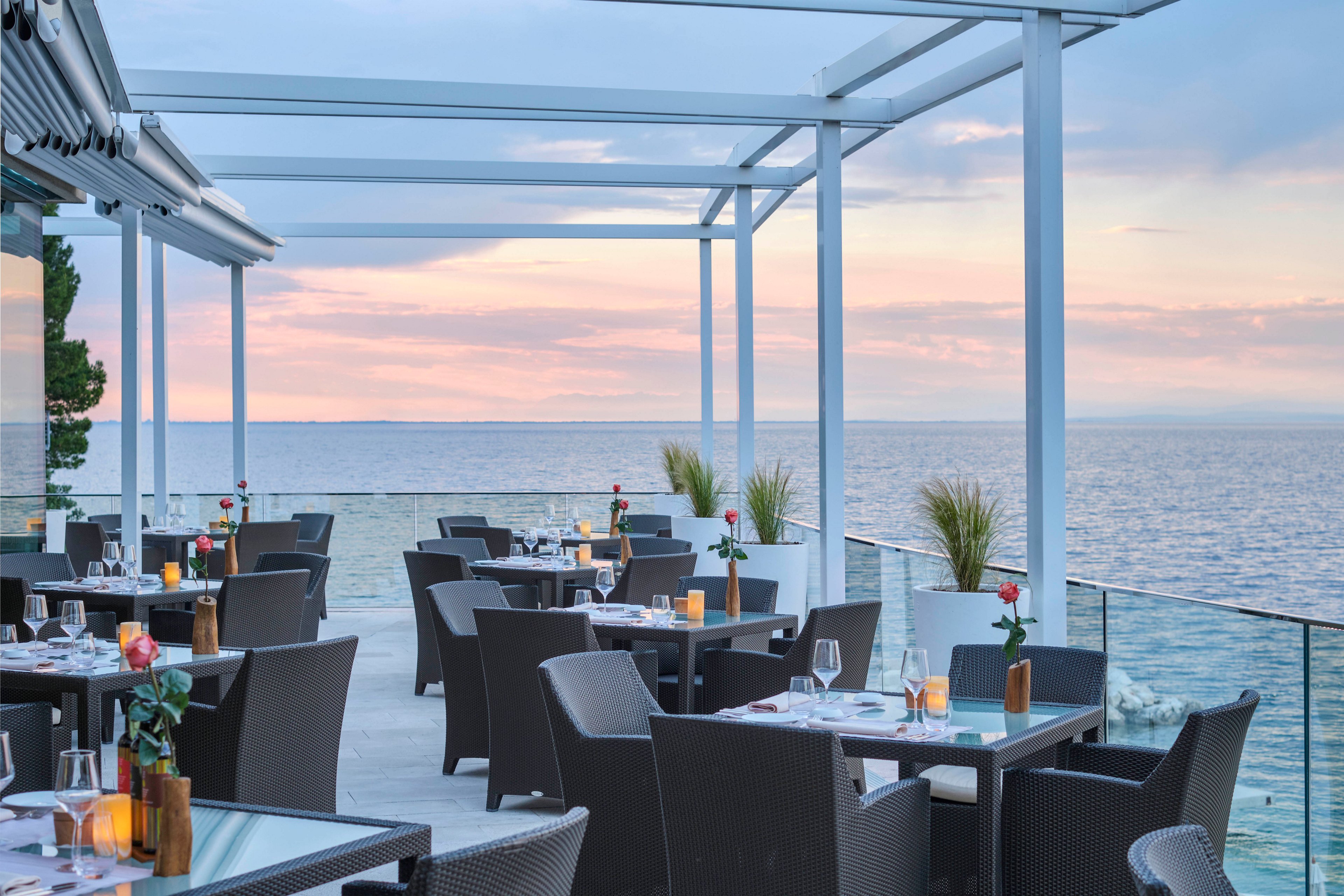 An upscale outdoor dining area overlooks a calm sea under a pastel sunset sky. Modern black chairs and tables are set with candles, glassware, and single red roses, creating a romantic and serene ambiance. The terrace features white pergolas and potted plants, with no visible people present. The tranquil setting highlights the beauty of waterfront dining and relaxed luxury.