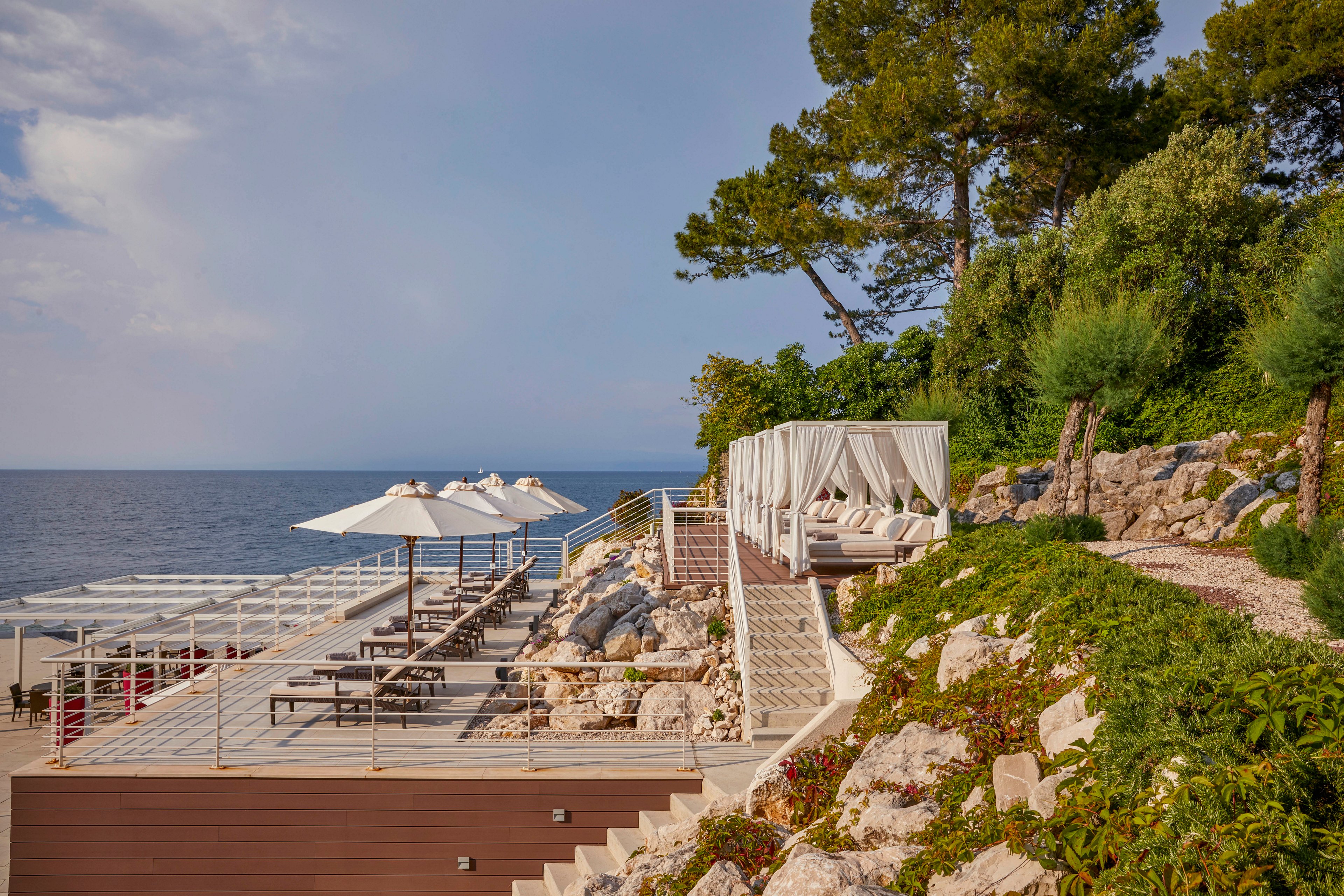 A stylish outdoor lounge area overlooks the sea, featuring shaded cabanas and rows of sun loungers with white umbrellas. The setting is built into a rocky hillside with lush greenery and stone steps leading down to the water. The atmosphere is tranquil and inviting, with soft neutral tones and a relaxed coastal vibe. No visible text or numbers are present in the image.
