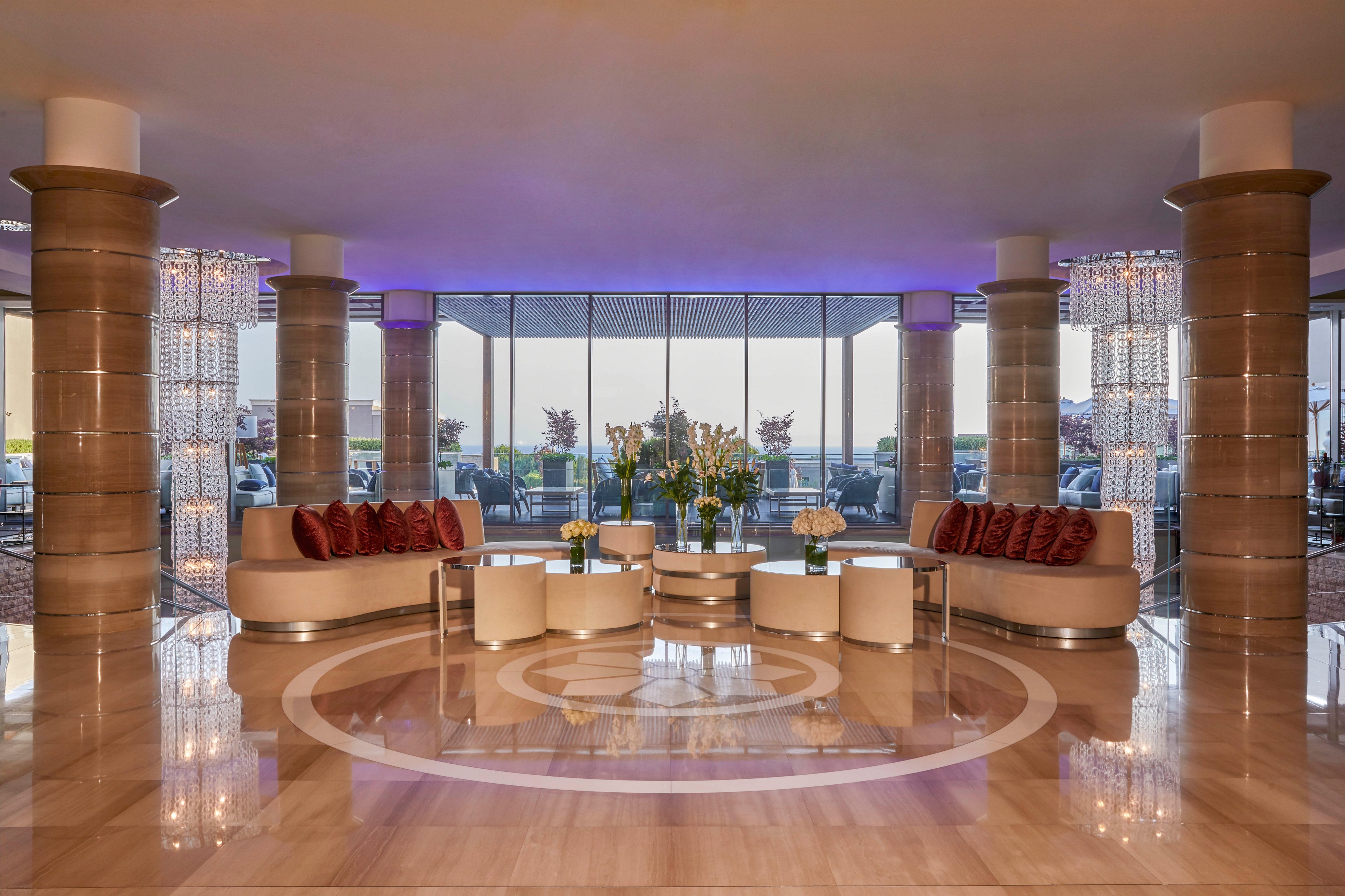 A luxurious hotel lobby features a symmetrical arrangement of curved sofas and round tables centered on a polished marble floor. Large columns and decorative lighting elements frame the space, while floor-to-ceiling windows reveal an outdoor terrace. The color palette is neutral with warm beige and soft purple lighting accents, creating a sophisticated and welcoming atmosphere. Fresh floral arrangements add a touch of nature to the contemporary setting.