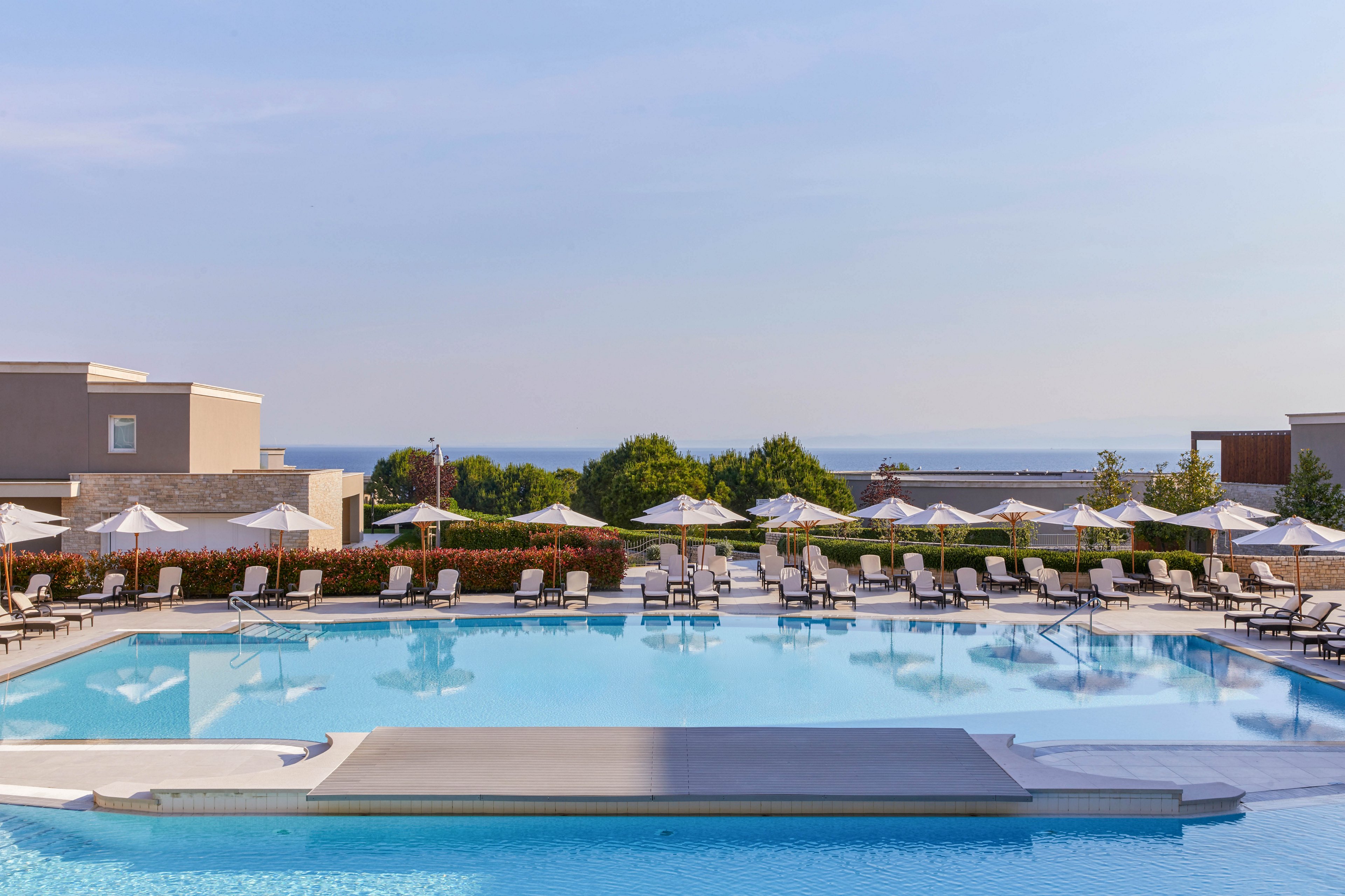 A large, tranquil swimming pool is the main focus, surrounded by numerous sun loungers and white umbrellas. The setting is an upscale resort with modern architecture, lush greenery, and a distant view of the sea under a clear blue sky. The scene is calm and inviting, ideal for relaxation and leisure activities.