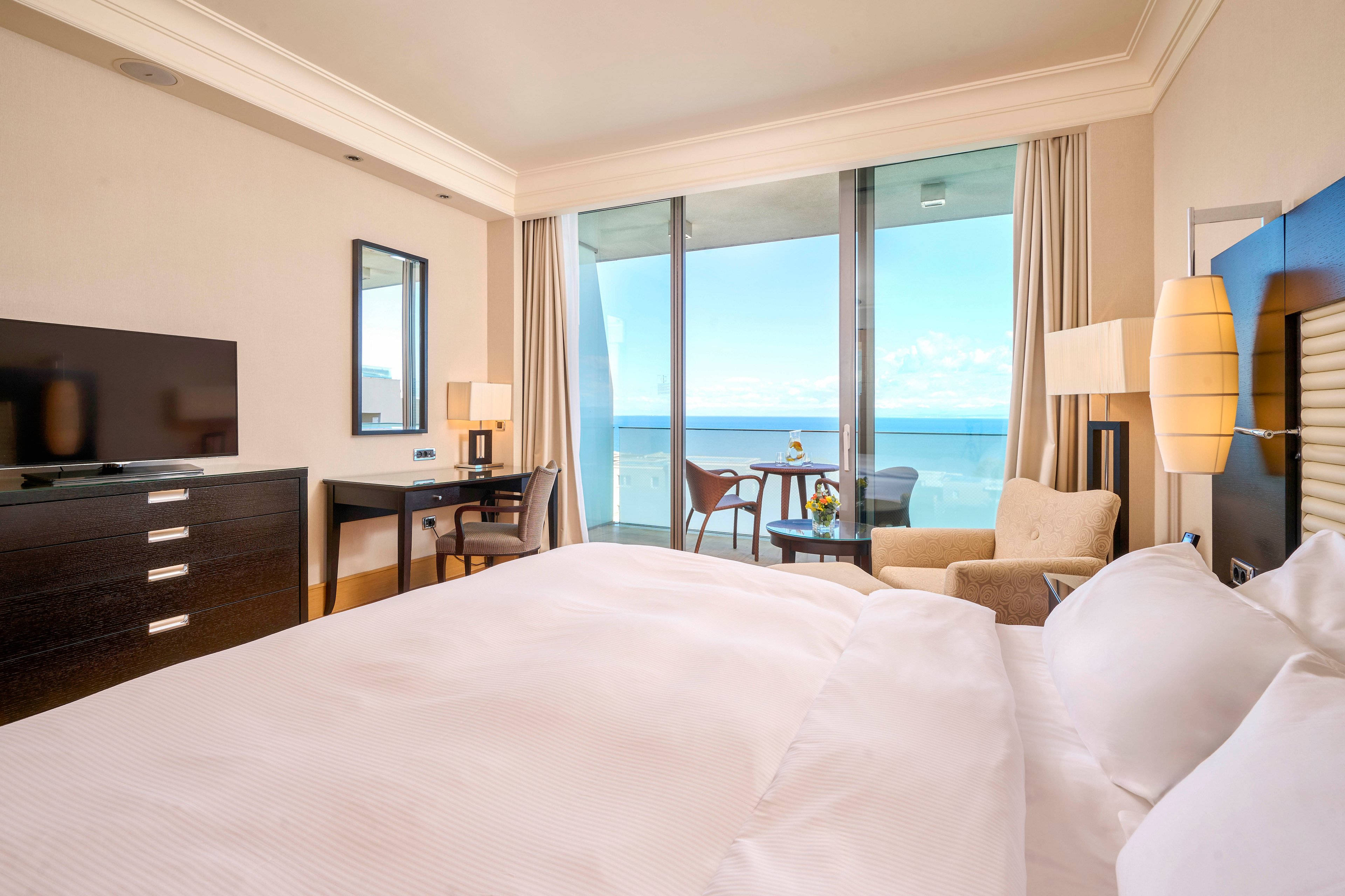 A spacious hotel room features a large bed with crisp white linens, a flat-screen television, and a work desk. The room opens to a balcony with seating, offering a clear view of the ocean and blue sky. Neutral tones and natural light create a calm, inviting atmosphere, while contemporary furnishings add a touch of elegance. No visible text or numbers are present in the image.