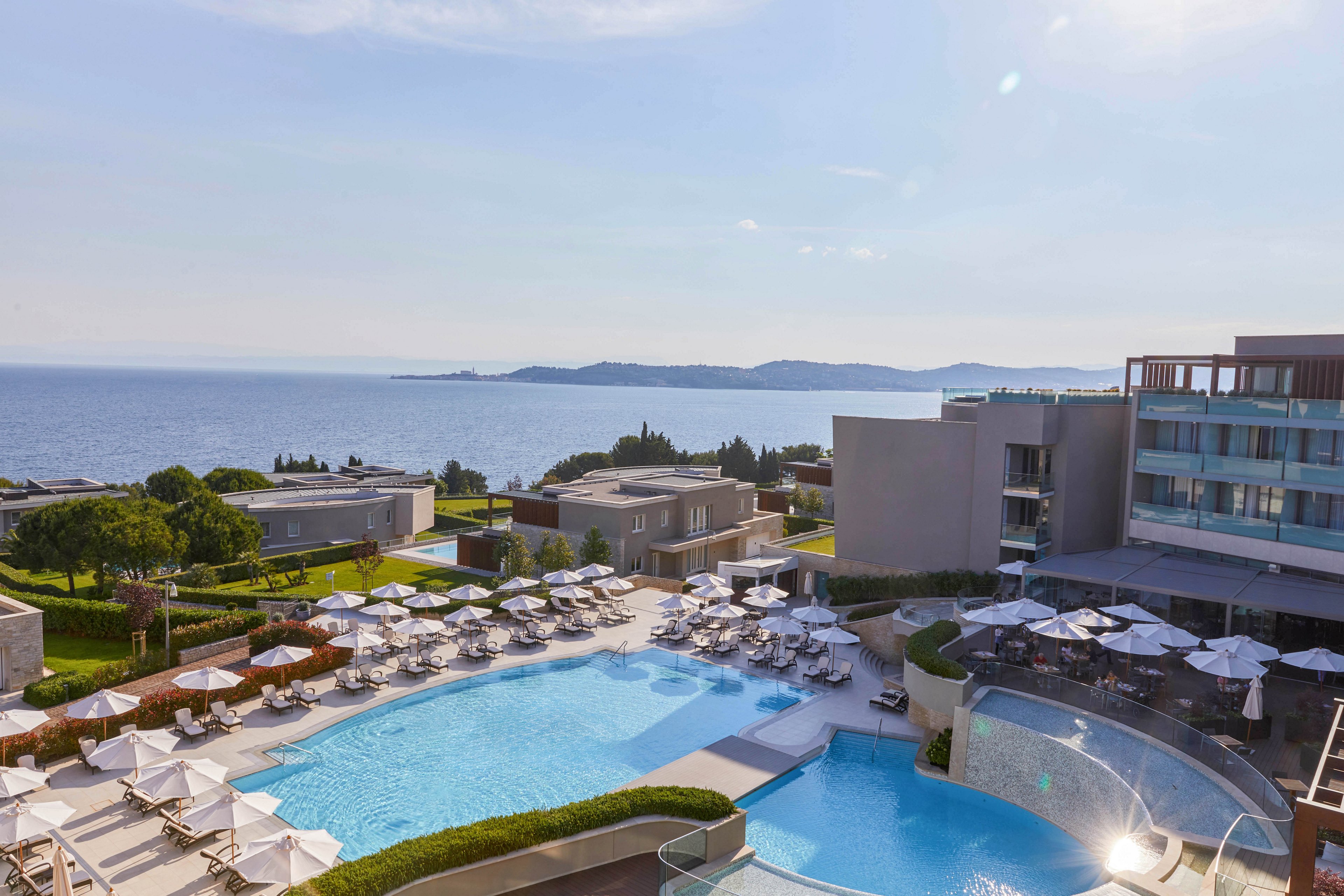 A modern resort features a large outdoor swimming pool surrounded by sun loungers and umbrellas. The setting overlooks a scenic coastline with calm blue sea and distant hills under a clear sky. The architecture is contemporary, with multiple buildings and landscaped greenery. The mood is tranquil and inviting, ideal for relaxation and leisure. No visible text or numeric values are present in the image.