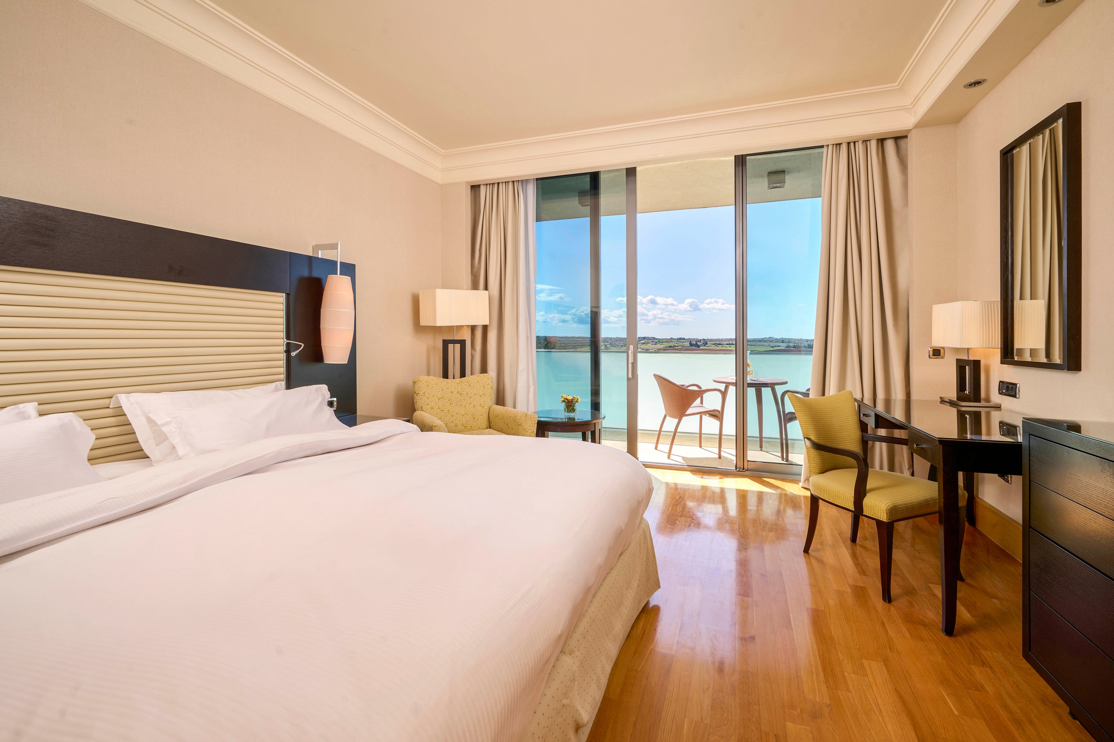 A spacious hotel room features a large bed with crisp white linens, a work desk, and a seating area. Floor-to-ceiling glass doors open to a balcony overlooking a serene body of water and blue sky. The room is decorated in neutral tones with wooden flooring, contemporary furniture, and soft lighting, creating a calm and inviting atmosphere. No visible text or numbers are present in the image.