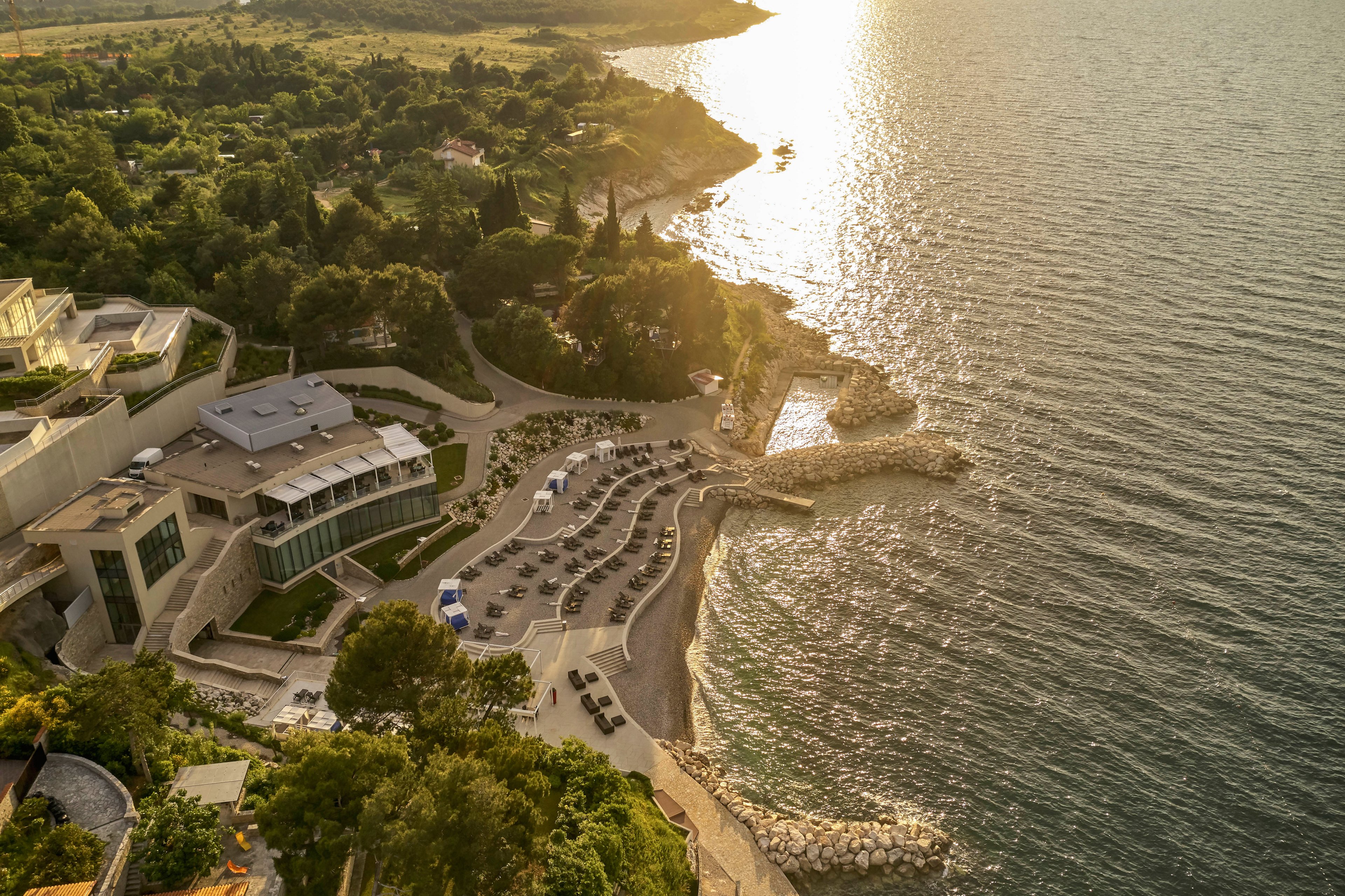 This image captures a modern seaside resort with terraced sun loungers and umbrellas along a rocky coastline. The setting sun casts a golden glow over the water and the lush greenery surrounding the property. The architecture features clean lines and large windows, blending with the natural landscape. The tranquil atmosphere suggests relaxation and leisure by the sea.
