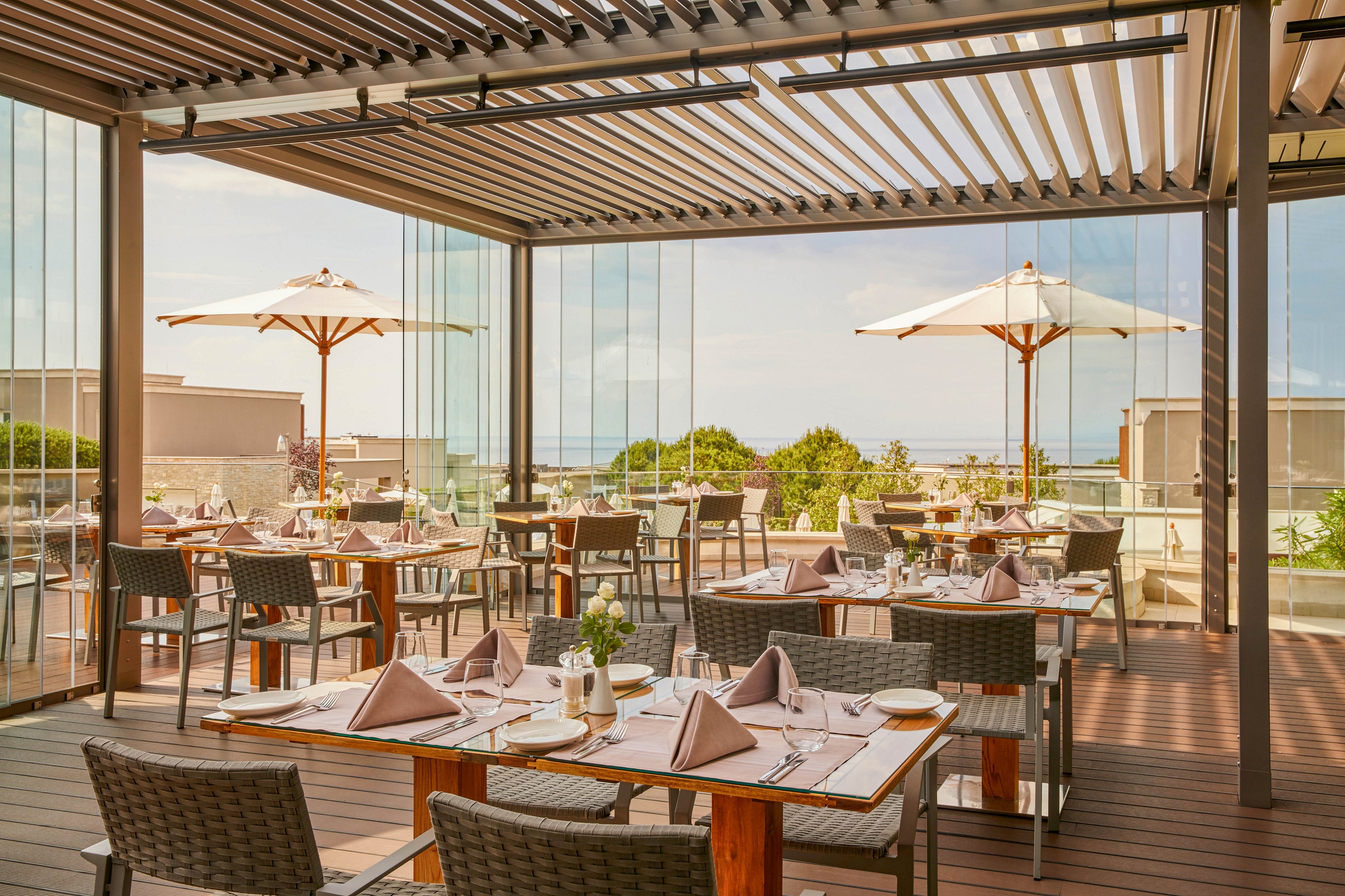 A modern outdoor restaurant terrace is set for dining, featuring neatly arranged tables with folded napkins and glassware. The space is covered by a stylish pergola and surrounded by glass walls, offering a clear view of the ocean and greenery. Large white umbrellas provide shade, and the overall atmosphere is calm and inviting with neutral tones. The setting suggests a relaxed, upscale dining experience in a coastal location.