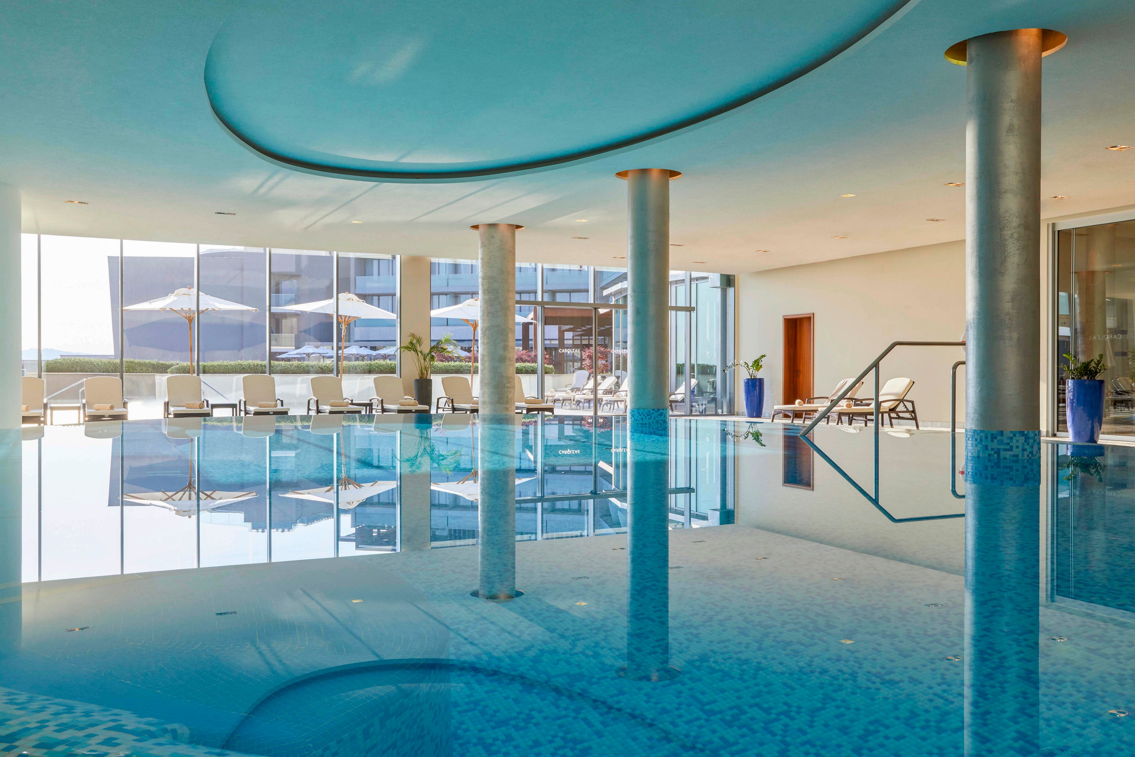 A spacious indoor swimming pool is featured in a contemporary setting with large floor-to-ceiling windows. Sun loungers and umbrellas are visible outside, creating a relaxing atmosphere. The pool area is accented by sleek columns and soft blue tones, with reflections enhancing the tranquil mood. No visible text or numbers are present in the image.