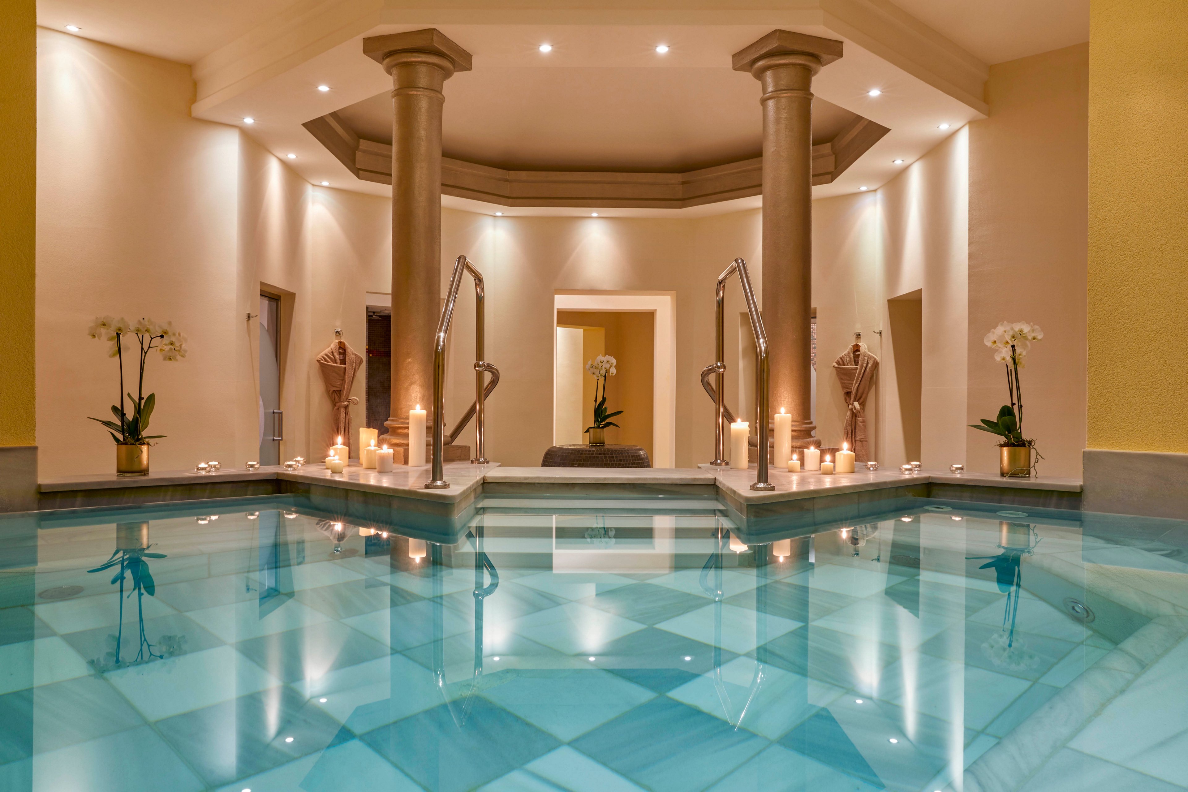 A serene indoor spa pool is featured, surrounded by elegant columns and soft, warm lighting. Numerous candles are placed around the pool, creating a tranquil and relaxing atmosphere. The water reflects the architectural details and the gentle glow of the candles. Orchids in pots add a touch of natural beauty to the sophisticated setting. No visible text or numbers are present in the image.