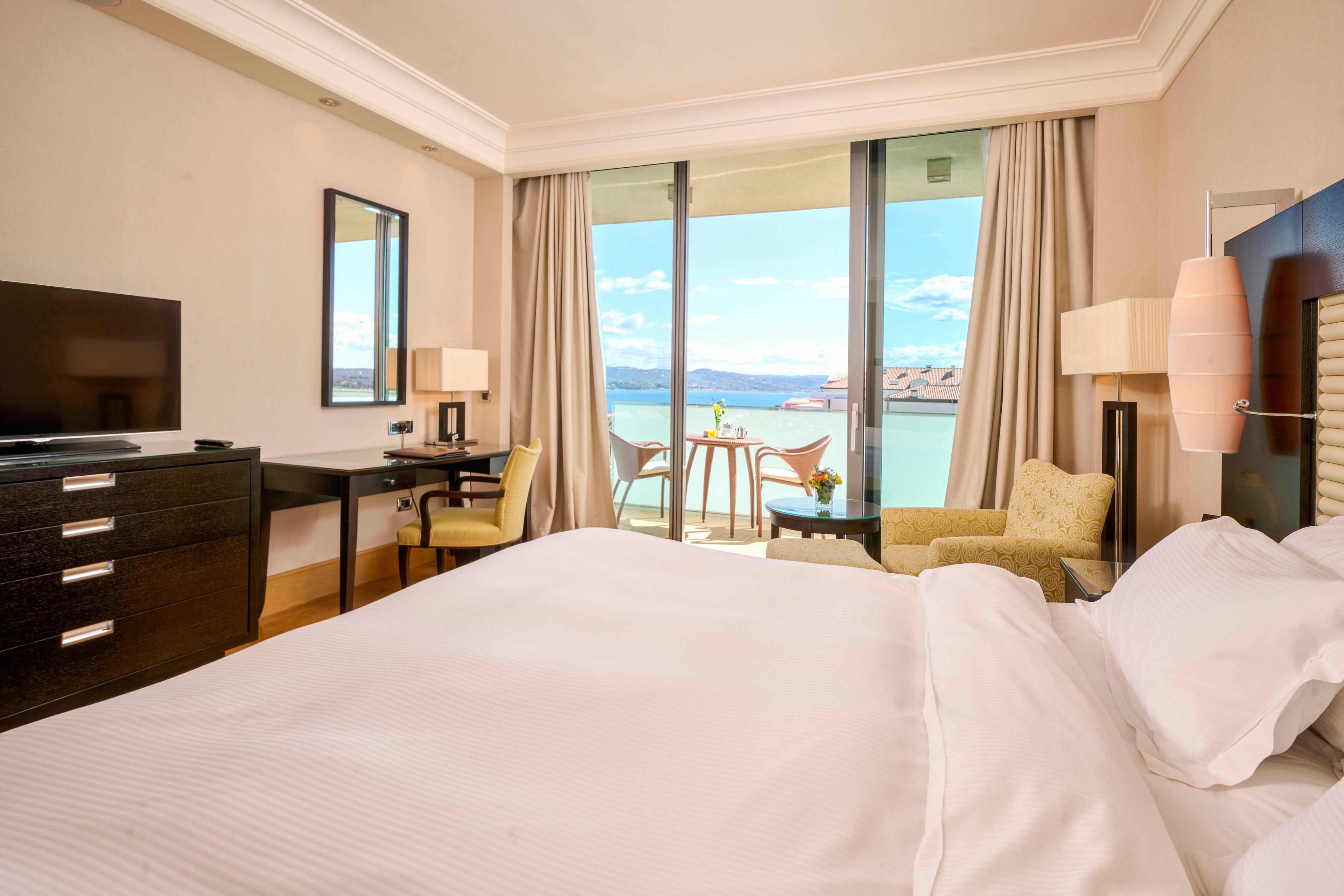 A spacious hotel room features a large bed with crisp white linens, a flat-screen TV, and a work desk. The room opens onto a balcony with a table and chairs, offering a scenic view of a calm body of water and distant mountains. The interior is decorated in neutral tones with soft lighting, creating a relaxing and inviting atmosphere. No visible text or numbers are present in the image.