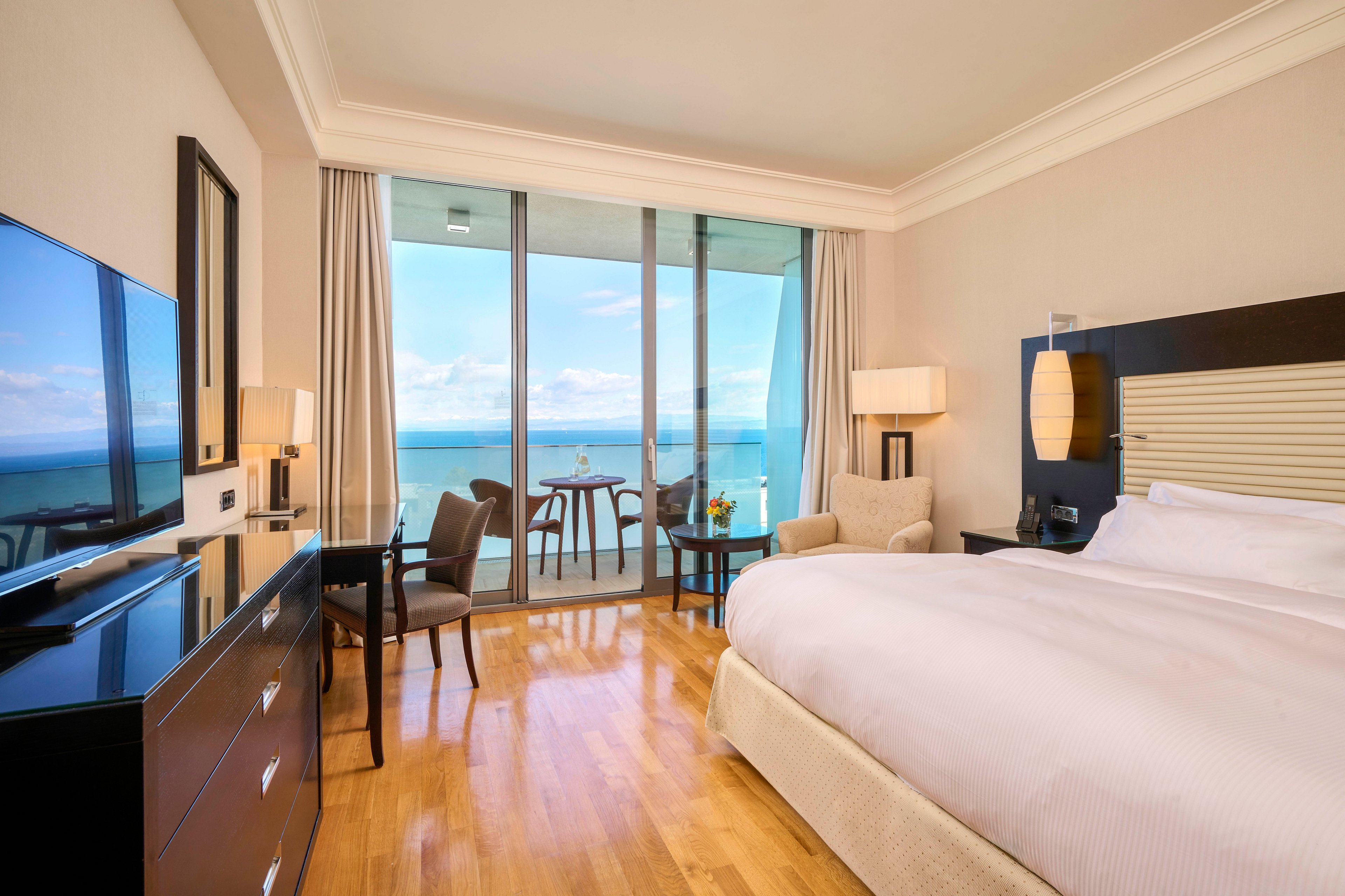 A spacious, contemporary hotel room features a large bed with crisp white linens, a flat-screen television, and elegant furnishings. Floor-to-ceiling glass doors open to a private balcony with a table and chairs, overlooking a serene ocean view. The room is decorated in neutral tones with wood flooring, creating a calm and inviting atmosphere. Natural light fills the space, highlighting the clean and modern design.