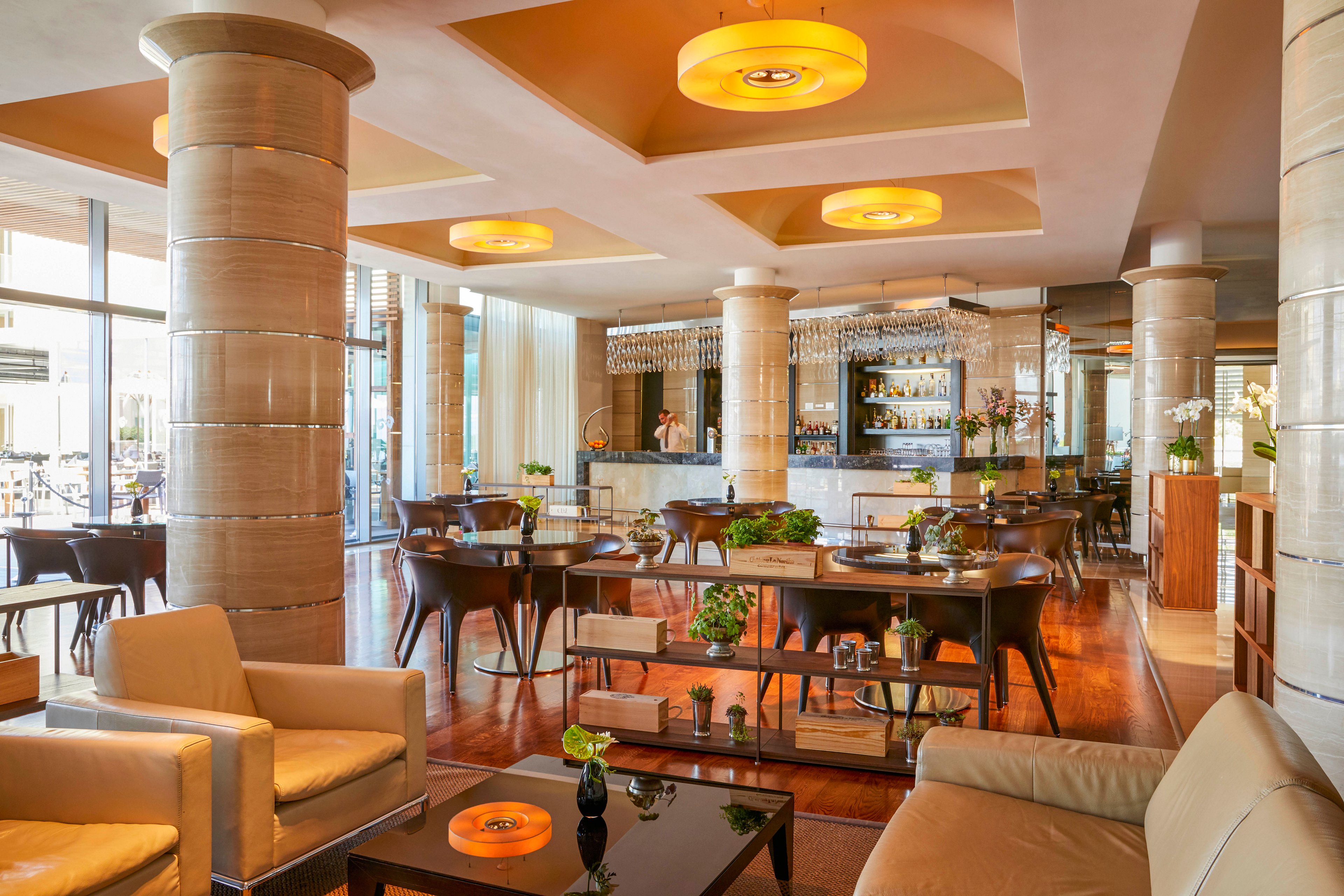 A spacious hotel restaurant lounge features contemporary furniture and large cylindrical columns. The setting includes a mix of dining tables and comfortable lounge chairs, with warm lighting fixtures and decorative plants enhancing the inviting atmosphere. The bar area is visible in the background, and the overall palette is neutral with wood and beige tones. No visible text or numbers are present in the image.