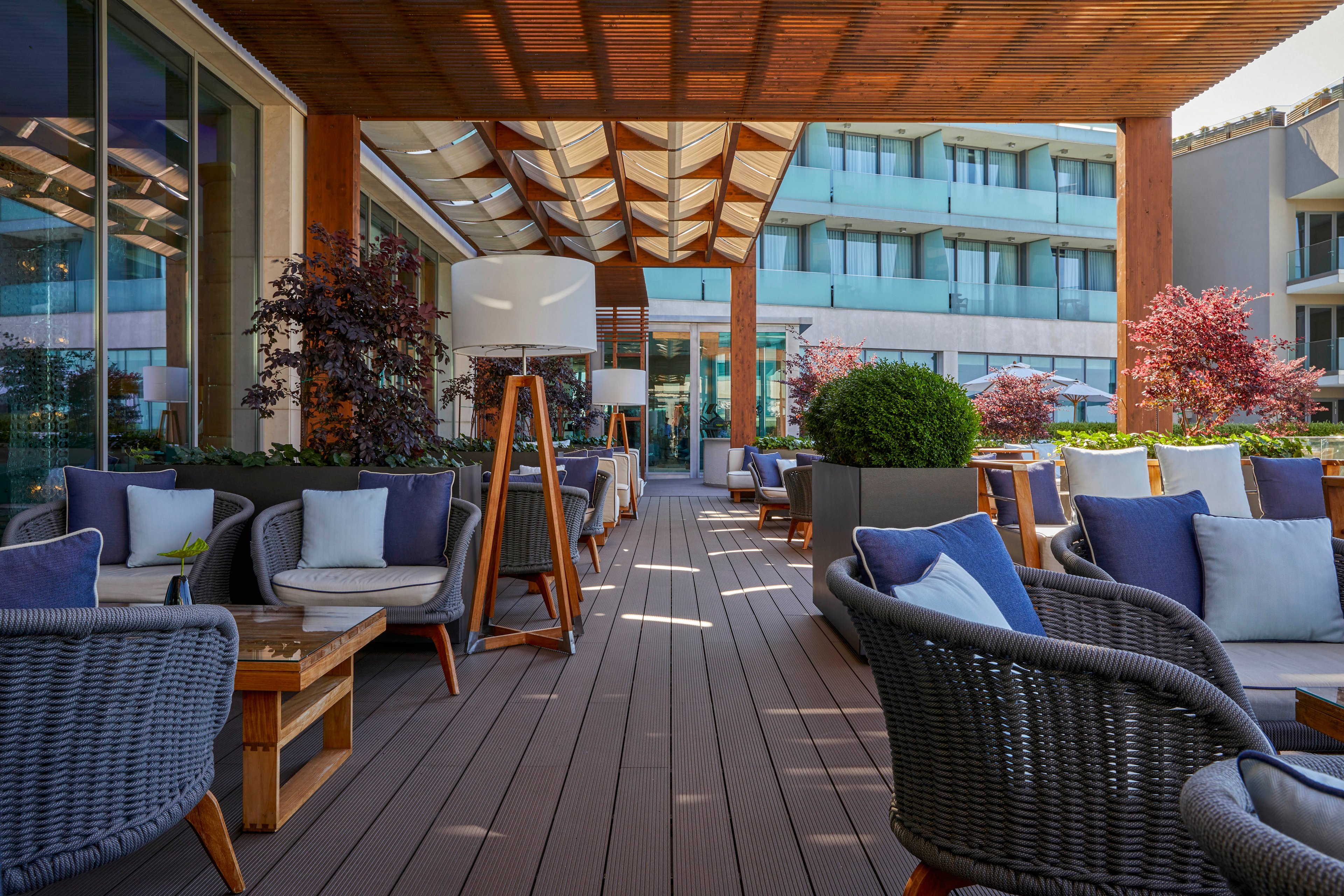A contemporary outdoor lounge features woven chairs and cushioned sofas arranged under a wooden pergola. The setting is part of a hotel or resort terrace, with large windows and manicured plants enhancing the relaxed atmosphere. Neutral tones and blue accents create a calm, inviting mood. No people are present, highlighting the tranquil environment. No visible text or numbers appear in the image.