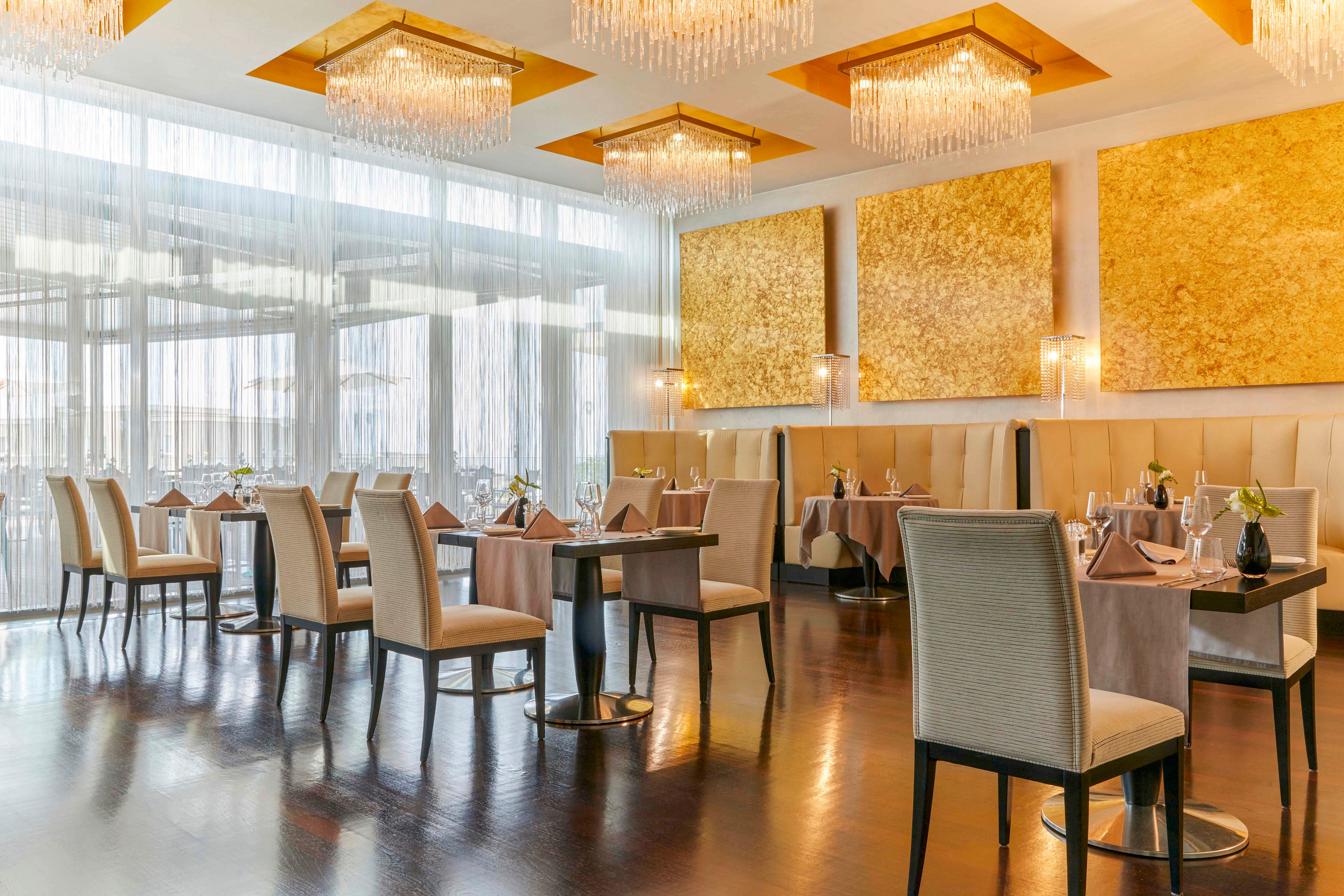 A sophisticated restaurant dining area features plush beige chairs and dark wooden tables, set with folded napkins and glassware. Large golden textured wall panels and crystal chandeliers add a luxurious touch to the contemporary space. Soft natural light filters through sheer curtains, creating a warm and inviting atmosphere. The polished dark floor reflects the ambient lighting, enhancing the upscale mood.