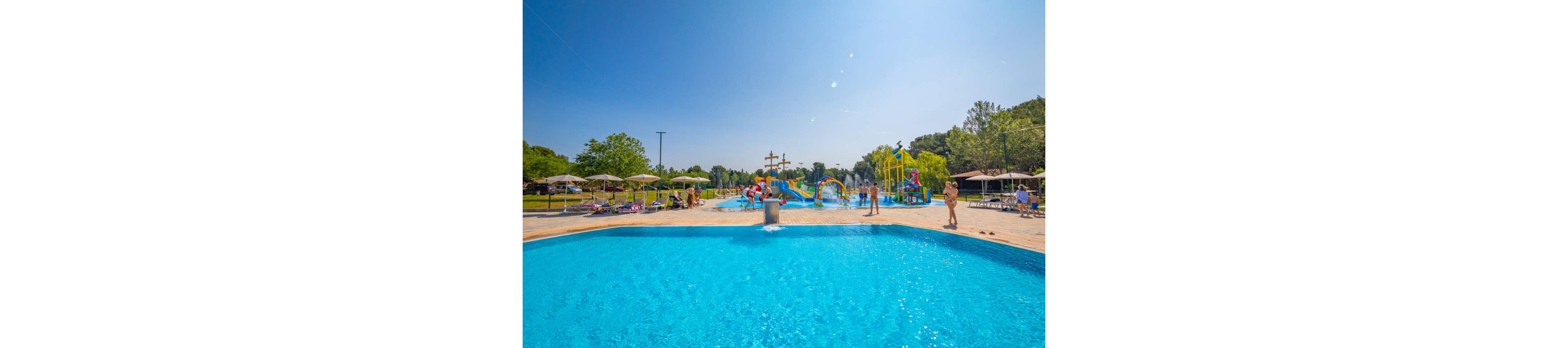 A bright blue outdoor swimming pool is the main focus of this sunny scene. In the background, families and children enjoy colorful water slides and splash structures. Sun loungers and umbrellas line the poolside area, with people relaxing and walking around. Green trees and a clear blue sky create a cheerful summer atmosphere.
