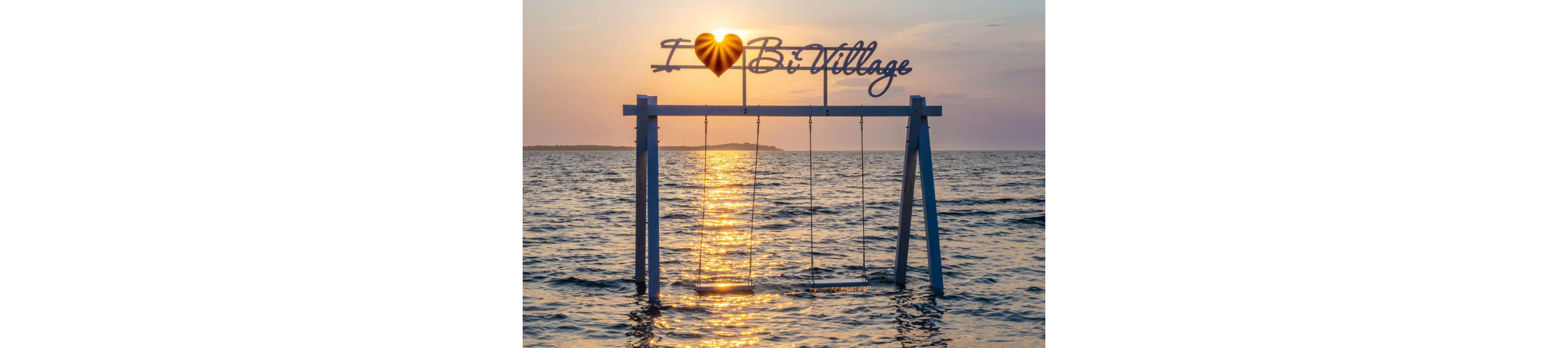 A pair of swings stands in shallow seawater beneath a decorative sign reading "I ♥ Bi Village" at sunset. The golden sun is low on the horizon, casting a bright reflection across the rippling water. Warm orange and pink tones fill the sky, contrasting with the cool blue sea. The scene feels tranquil and romantic, inviting relaxation and seaside enjoyment.