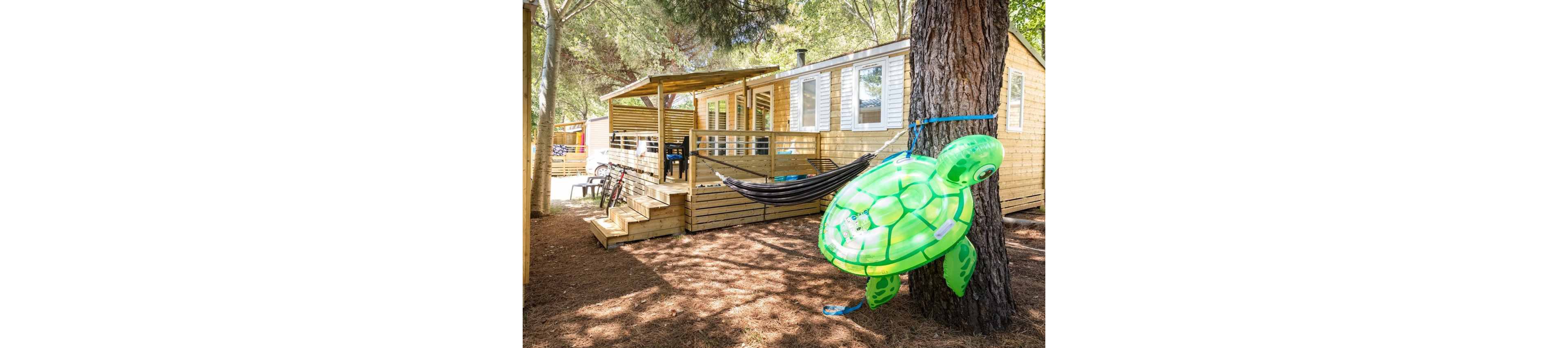 A wooden mobile home is set among tall trees in a shaded outdoor campsite. A bright green inflatable turtle float is tied to a tree in the foreground, adding a playful touch. A black hammock hangs between trees near the cabin’s deck, which is furnished with chairs and outdoor gear. Sunlight filters through the leaves, creating a relaxed vacation atmosphere in nature.