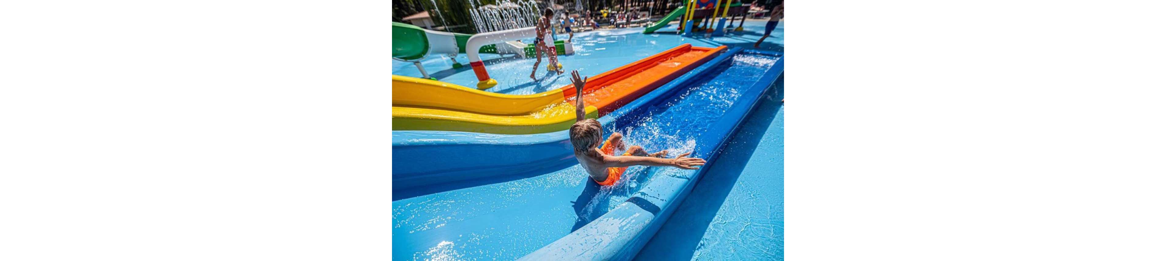 A young child slides down a bright blue water slide at an outdoor water park. Multiple colorful slides and shallow pools create a playful, family-friendly setting. Other children and adults are visible in the background enjoying fountains and water features. The scene is sunny and vibrant, emphasizing fun, motion, and summer recreation.