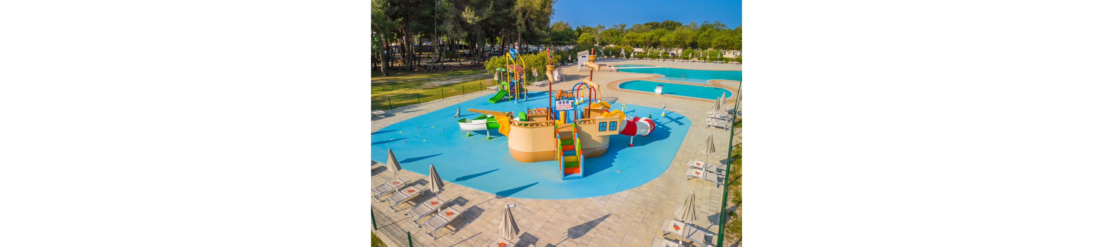 A brightly colored pirate ship themed water playground sits in the center of a shallow splash pool. The scene is captured from above, showing slides, climbing areas, and water features designed for children. Surrounding the play structure are sun loungers with closed umbrellas on a tiled deck. In the background, a larger swimming pool and a line of trees indicate a family-friendly resort or campground setting.
