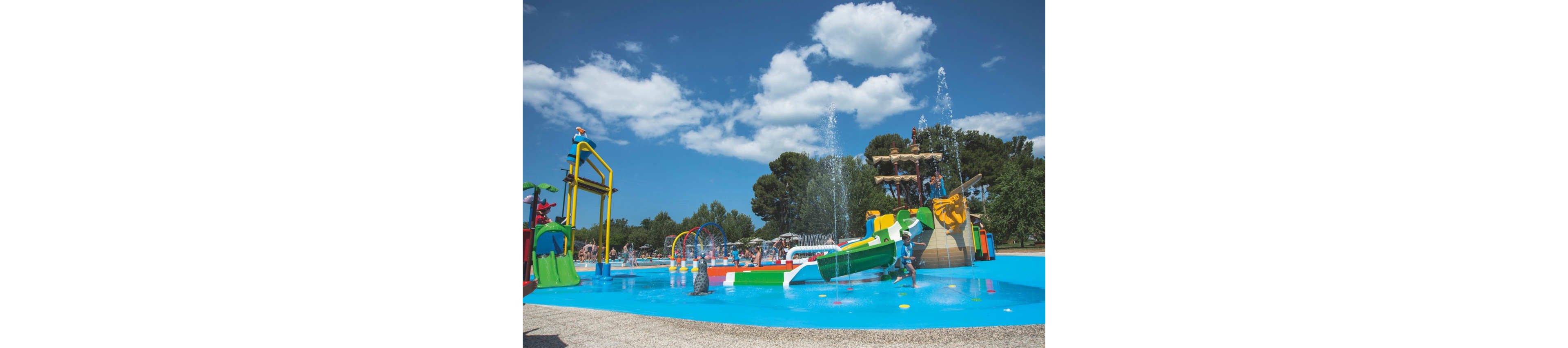A vibrant outdoor splash park features children playing on colorful water slides and climbing structures. Fountains shoot water into the bright blue splash pool under a sunny sky with scattered clouds. Adults and other children relax and watch in the background near trees and additional play equipment. The scene conveys a fun, family-friendly summer atmosphere in a recreational water playground.