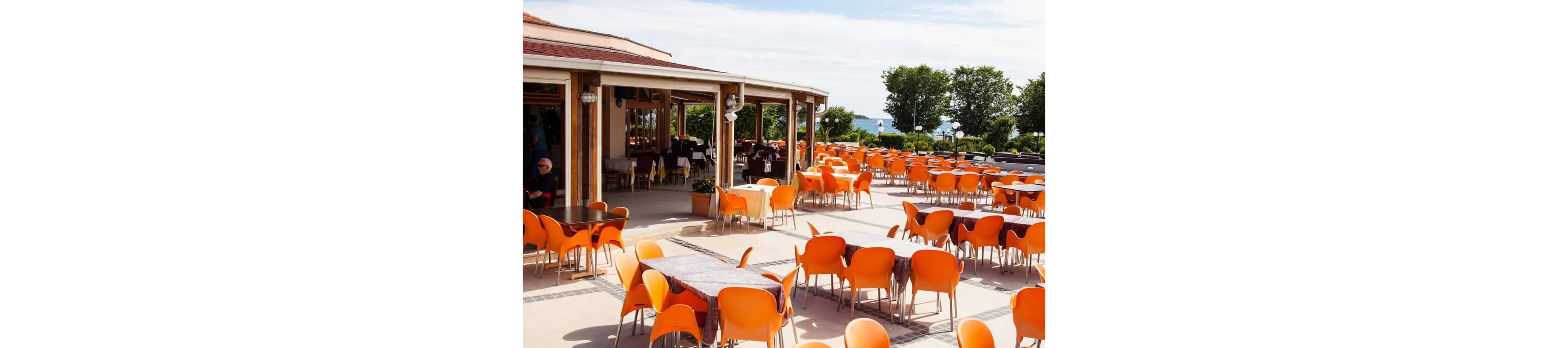An expansive outdoor café terrace is filled with rows of bright orange chairs and tables. The seating area extends from a covered patio attached to a building with large windows. Sunlight illuminates the open space, suggesting a warm and pleasant day. Green trees and distant landscape provide a natural backdrop, creating a relaxed and inviting atmosphere.