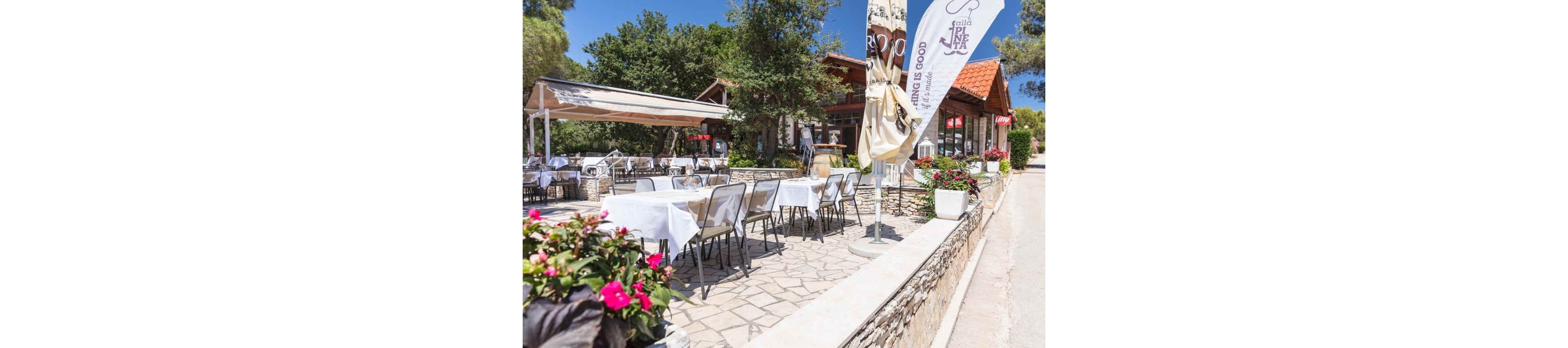 An inviting outdoor restaurant terrace is set with white tablecloths and empty chairs under a bright blue sky. The dining area is paved with stone and bordered by low stone walls and potted flowers. A building with a red-tiled roof and large windows stands behind the terrace, partially shaded by trees. Tall flags and umbrellas add vertical accents, while colorful blossoms in planters enhance the cheerful atmosphere.