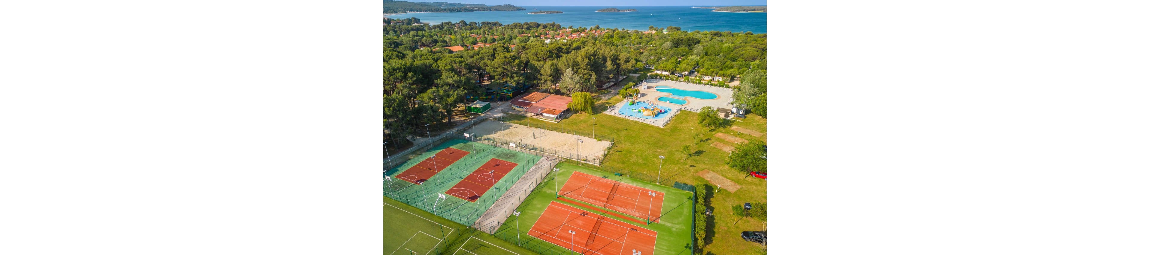 A large outdoor sports resort is captured from above, featuring multiple red tennis courts and a sand court. Nearby, a curving swimming pool area with sun loungers is surrounded by trees and landscaped greenery. Residential buildings and forested areas stretch toward the coastline, with blue sea and small islands visible in the distance. The scene suggests an active vacation destination combining recreation, relaxation, and scenic coastal views.