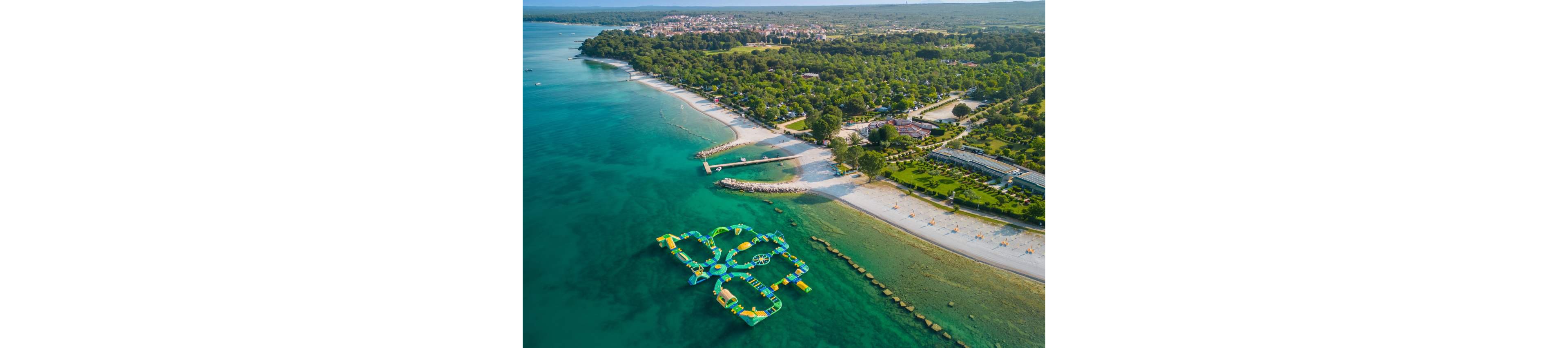 A colorful inflatable water park floats just offshore in clear turquoise water. The coastline features sandy beaches, piers, and neatly arranged sunbathing areas. Behind the shore, a lush green resort area with trees, paths, and buildings stretches toward a distant town. The scene suggests a popular summer holiday destination with calm seas and bright weather.