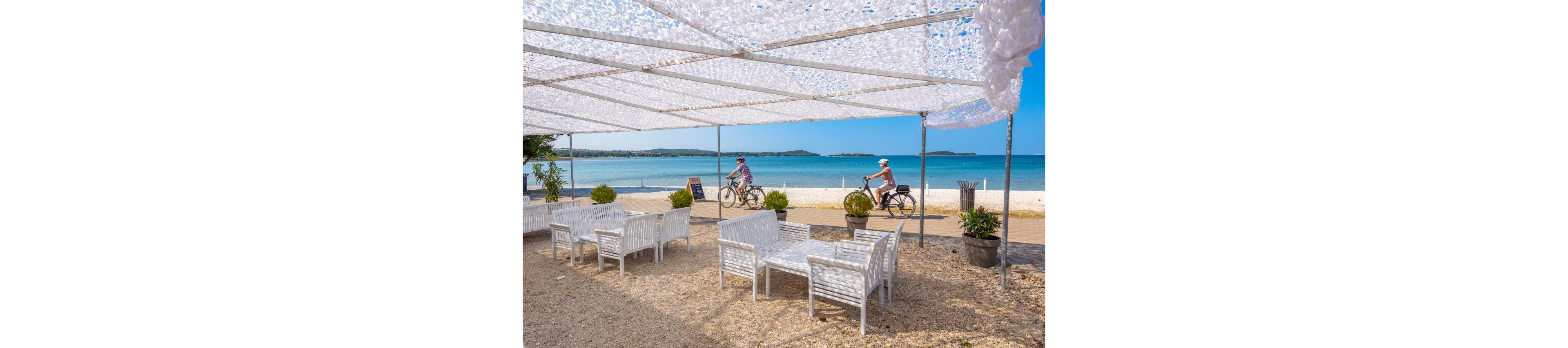 An open-air seaside café with white wooden tables and chairs sits under a decorative white canopy. In the background, two adults ride bicycles along a paved path beside the sandy beach. The calm blue sea and distant shoreline create a relaxed coastal atmosphere. Potted plants and a small signboard add detail to the casual outdoor setting.