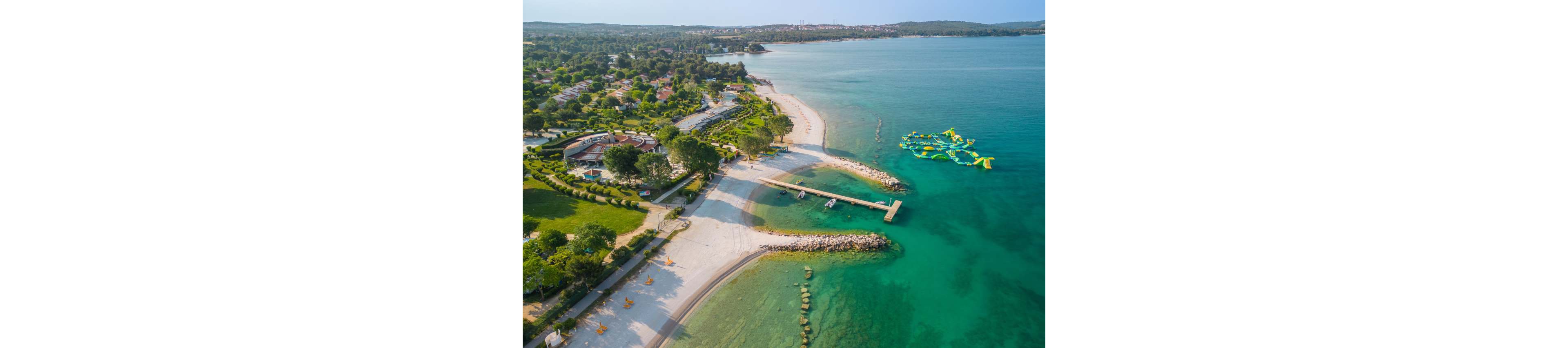 A coastal resort stretches along a curved sandy beach beside clear turquoise water. The aerial perspective reveals landscaped gardens, walking paths, and several buildings near the shoreline. A long pier extends into the sea, with small boats moored nearby. Off the coast, a floating inflatable water park adds a playful element to the tranquil seaside scene.
