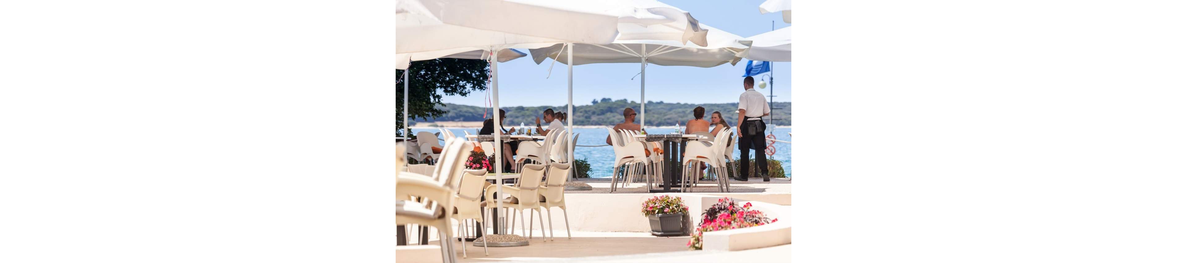 An outdoor café terrace overlooks a calm seaside landscape under large white umbrellas. Several adults sit at tables enjoying food and drinks while a waiter attends to one of the groups. Stacks of empty chairs and potted flowers decorate the bright, sunlit patio. The distant shoreline and blue water create a relaxed holiday atmosphere. The overall scene suggests leisurely dining in a coastal resort setting.