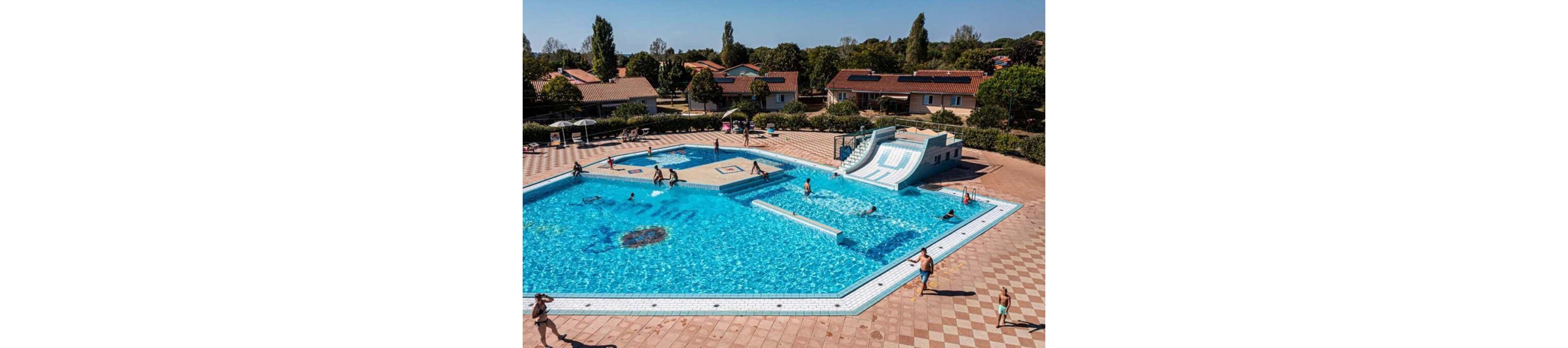 An outdoor swimming pool at a holiday resort is filled with people enjoying the water. Children and adults play near the built-in water slides and shallow areas. The pool is surrounded by a wide tiled deck where several people walk or relax. In the background, low houses, trees, and clear skies create a sunny vacation atmosphere.