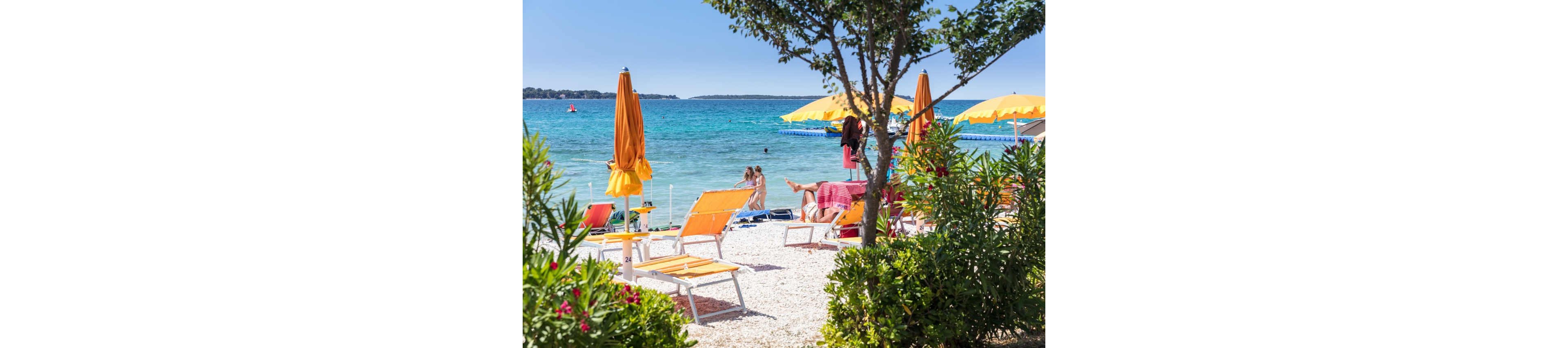 A bright seaside scene shows orange lounge chairs and umbrellas arranged on a white pebble beach. People stand and walk near the shoreline, enjoying the clear turquoise water. Yellow umbrellas line the water’s edge, and lush green plants frame the foreground. The overall atmosphere is relaxed, summery, and inviting for a coastal vacation.