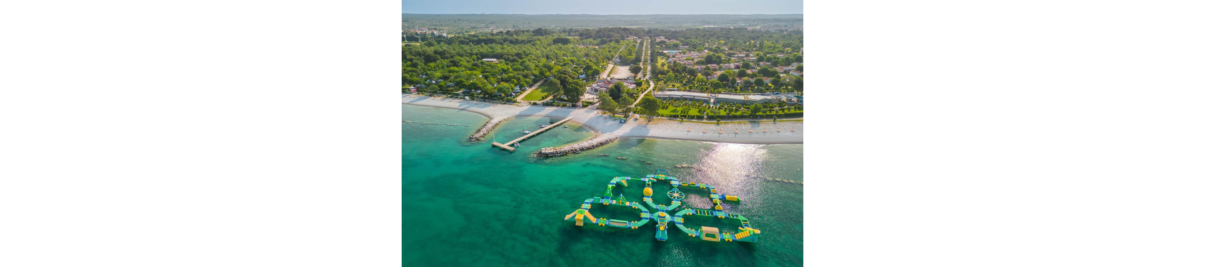 An aerial view shows a colorful inflatable water park floating on clear turquoise sea water near a sandy coastline. The floating structure features interconnected platforms, slides, and climbing elements in bright green and yellow tones. Along the shore, a long beach, piers, and breakwaters extend into the water, backed by trees and low-rise buildings. The scene suggests a popular seaside resort area with recreational facilities and calm, inviting waters.